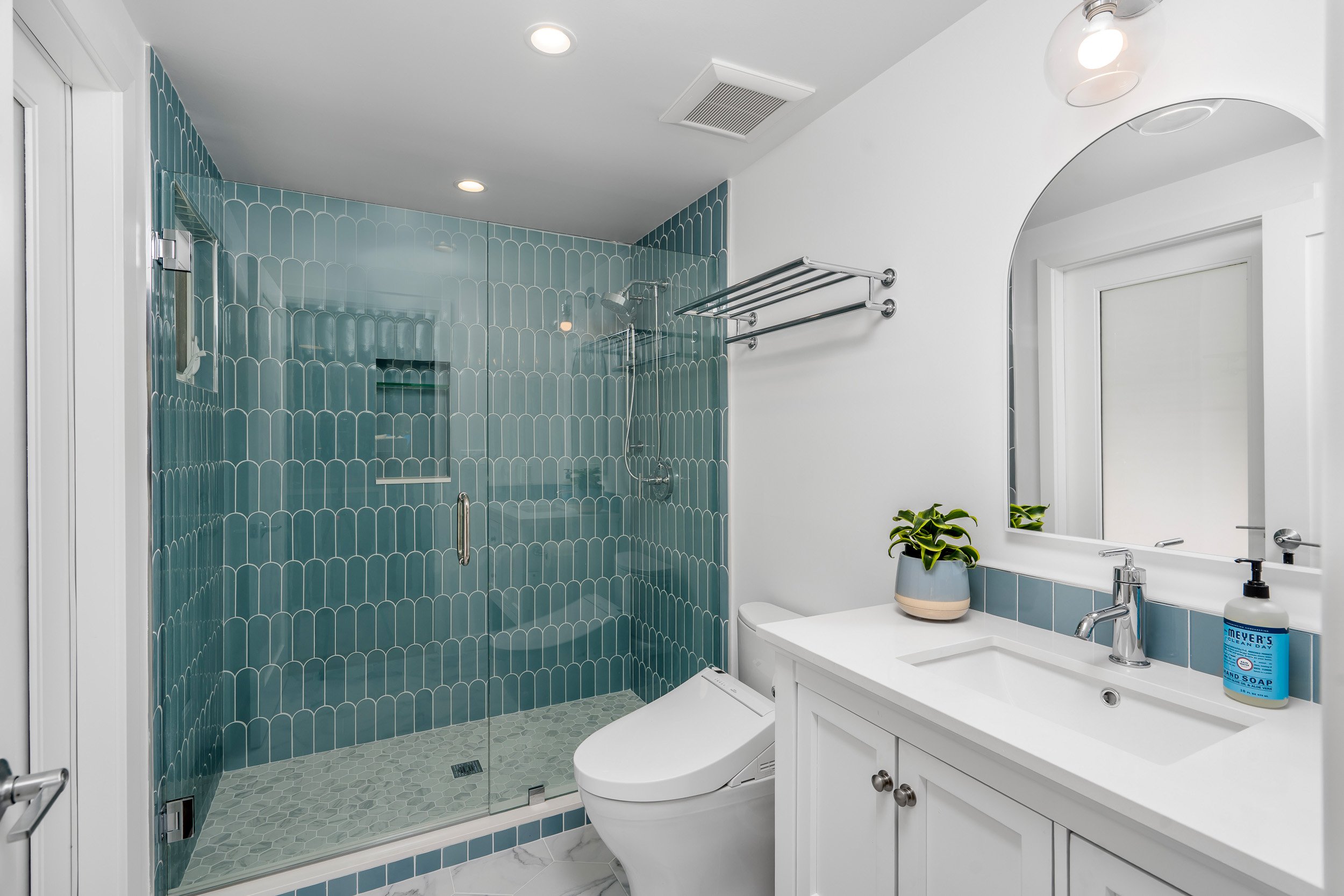 Bathroom with a glass shower enclosure, teal tile walls, white vanity with a mirror, potted plant, toilet, and essentials like hand soap and a bottle of hand sanitizer.