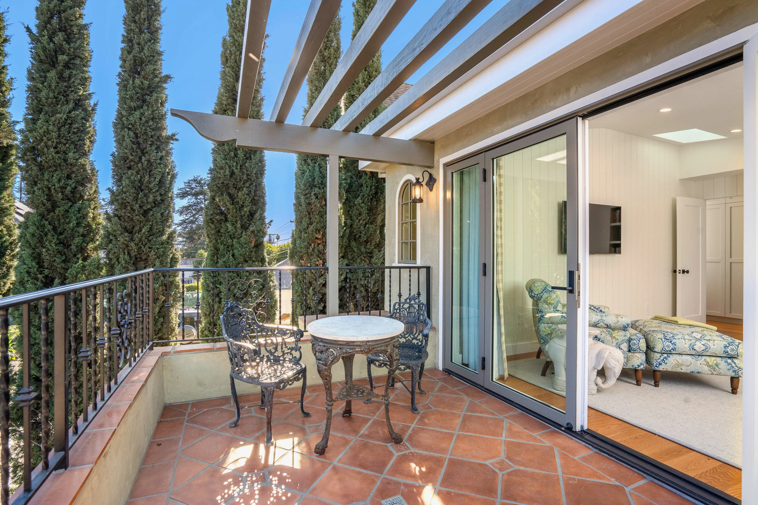 A balcony with a round marble table and metal chairs, overlooking tall cypress trees, with a sliding glass door leading to an interior living space.