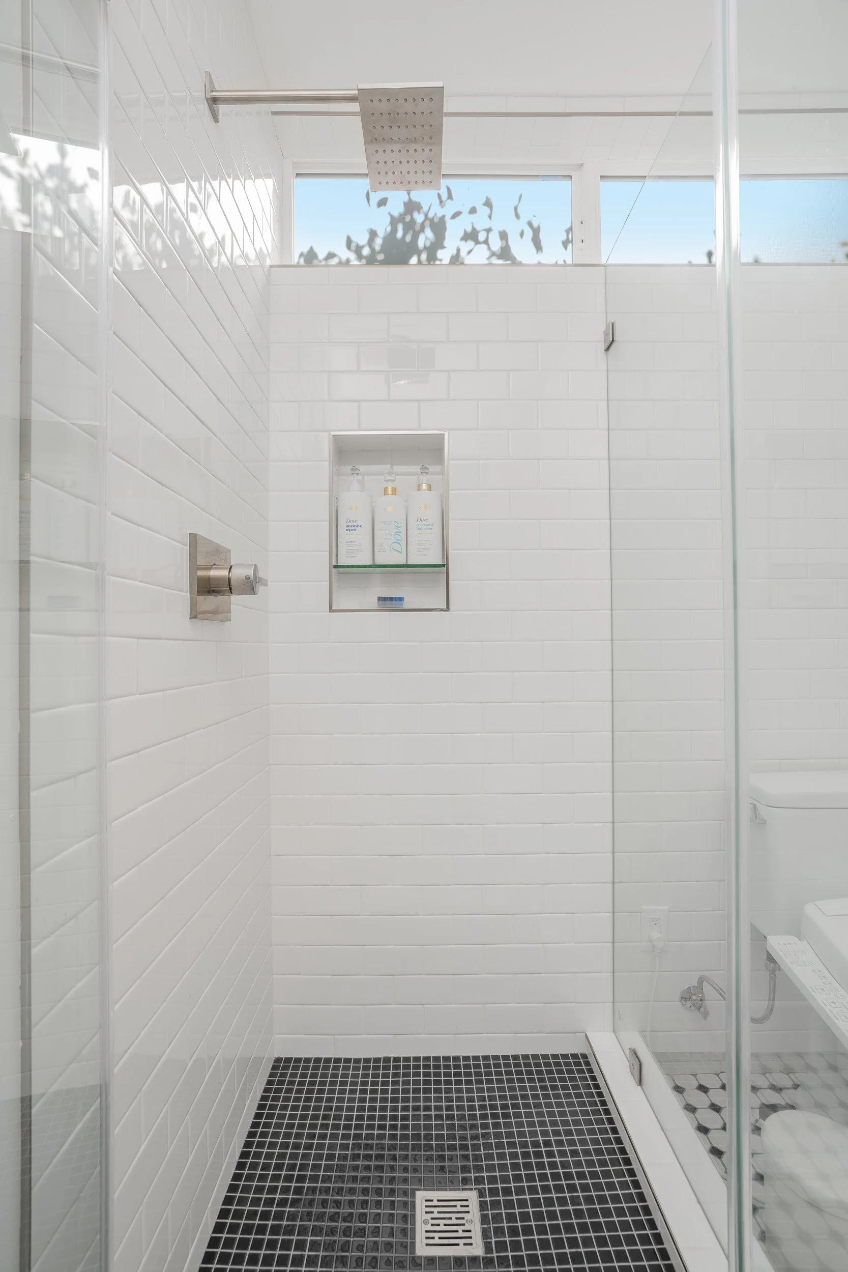 Clean modern shower with white subway tiles and black floor tiles, a built-in shelf with soap and shampoo bottles, a rainfall showerhead, and a window near the ceiling.