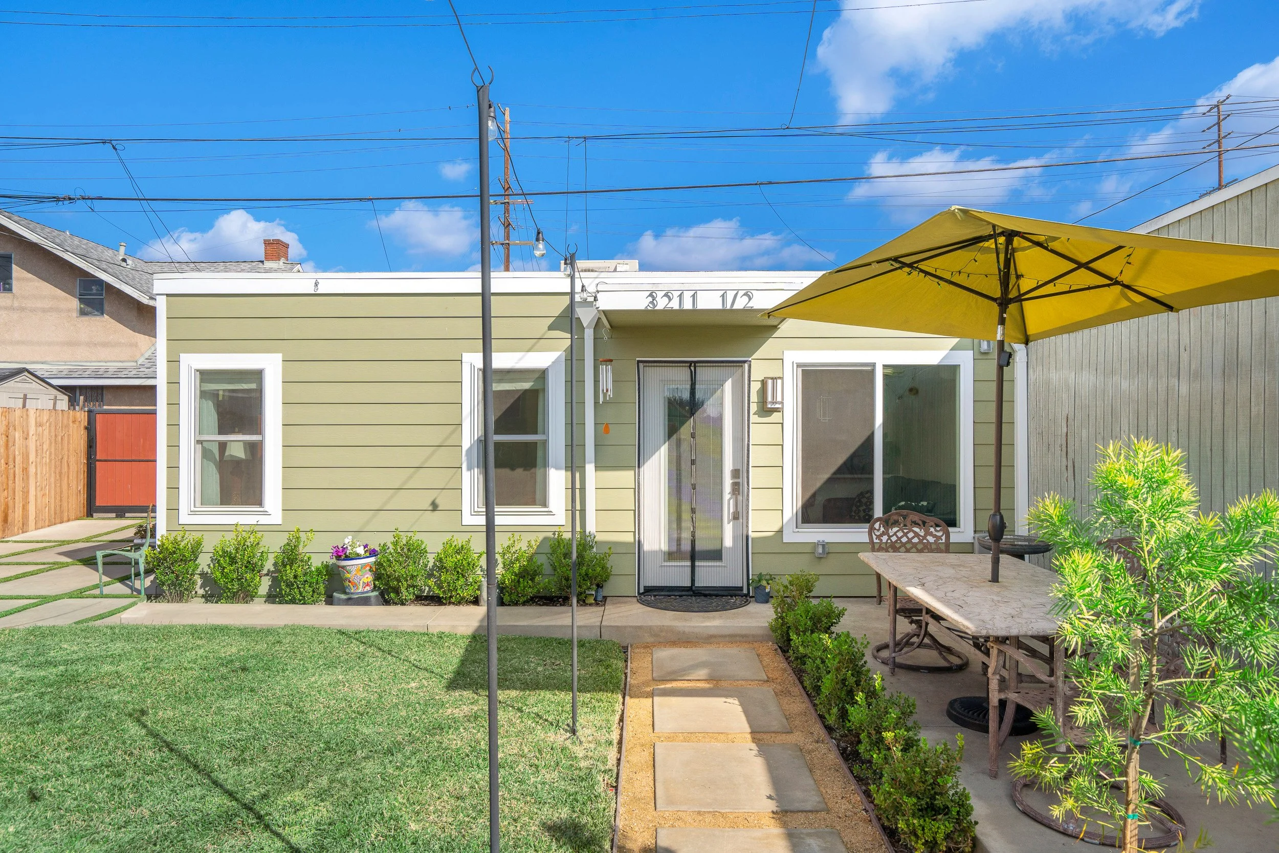 Backyard view of a yellow house with two windows and a glass door, surrounded by a well-kept lawn, potted plants, a concrete pathway, outdoor furniture, a yellow patio umbrella, and power lines overhead against a partly cloudy blue sky.