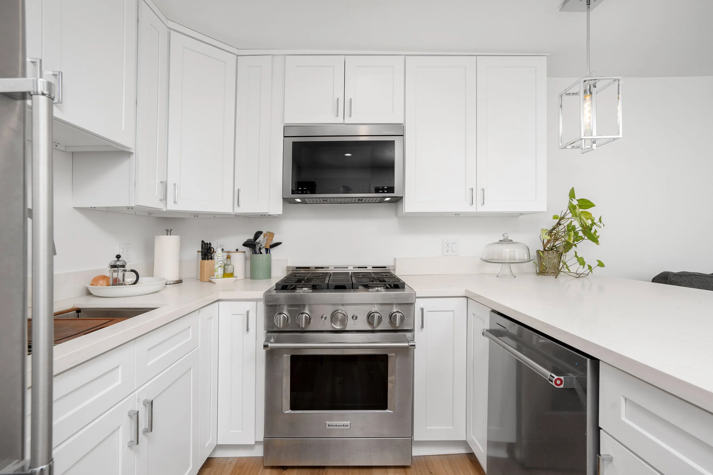 Modern kitchen with white cabinets, stainless steel appliances, a gas stove, and a microwave oven. A plant is on the right side of the counter, and there are kitchen utensils and containers on the left.