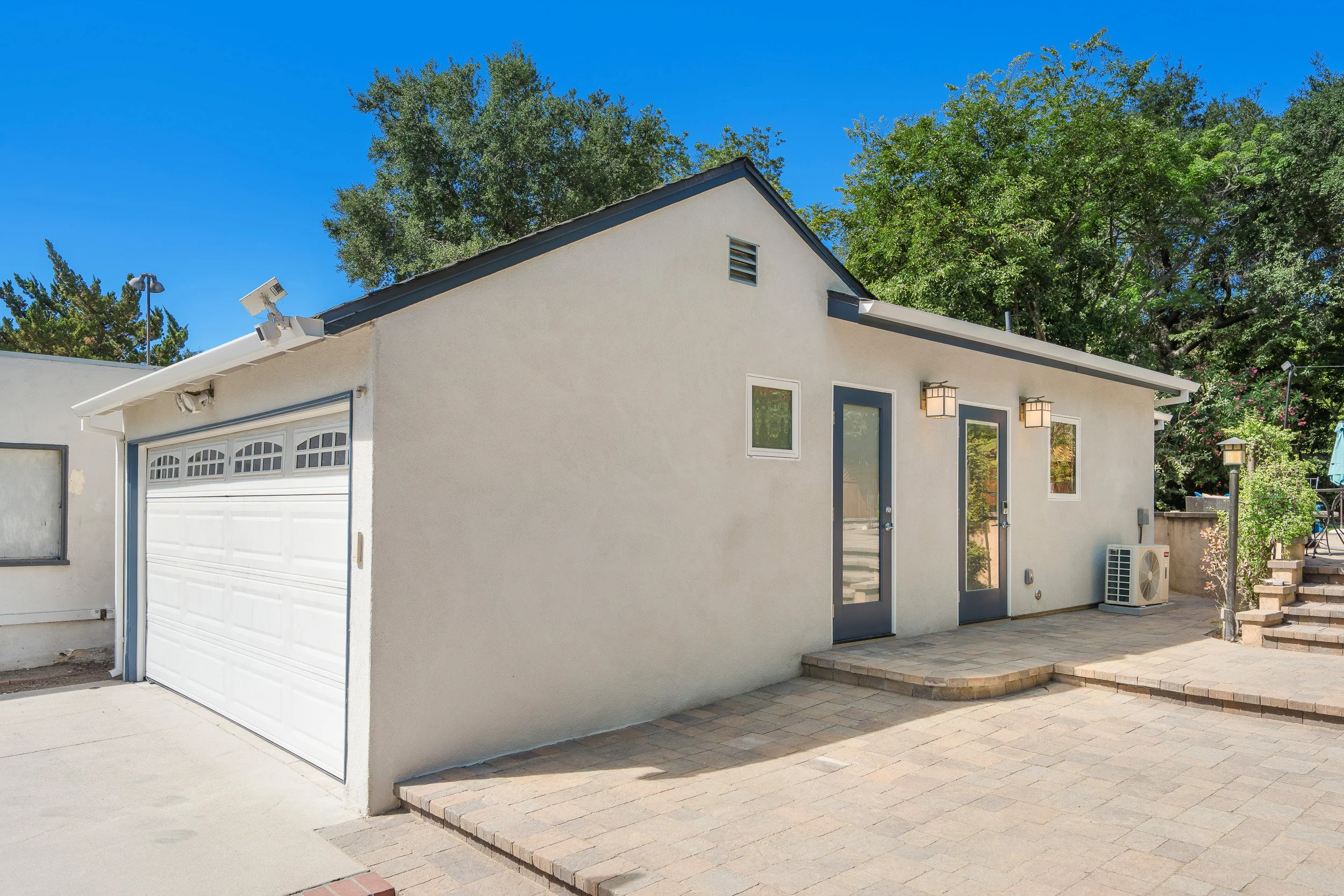 A modern, white exterior house with a two-car garage, blue front door, and a concrete patio with steps, surrounded by trees and a bright blue sky.
