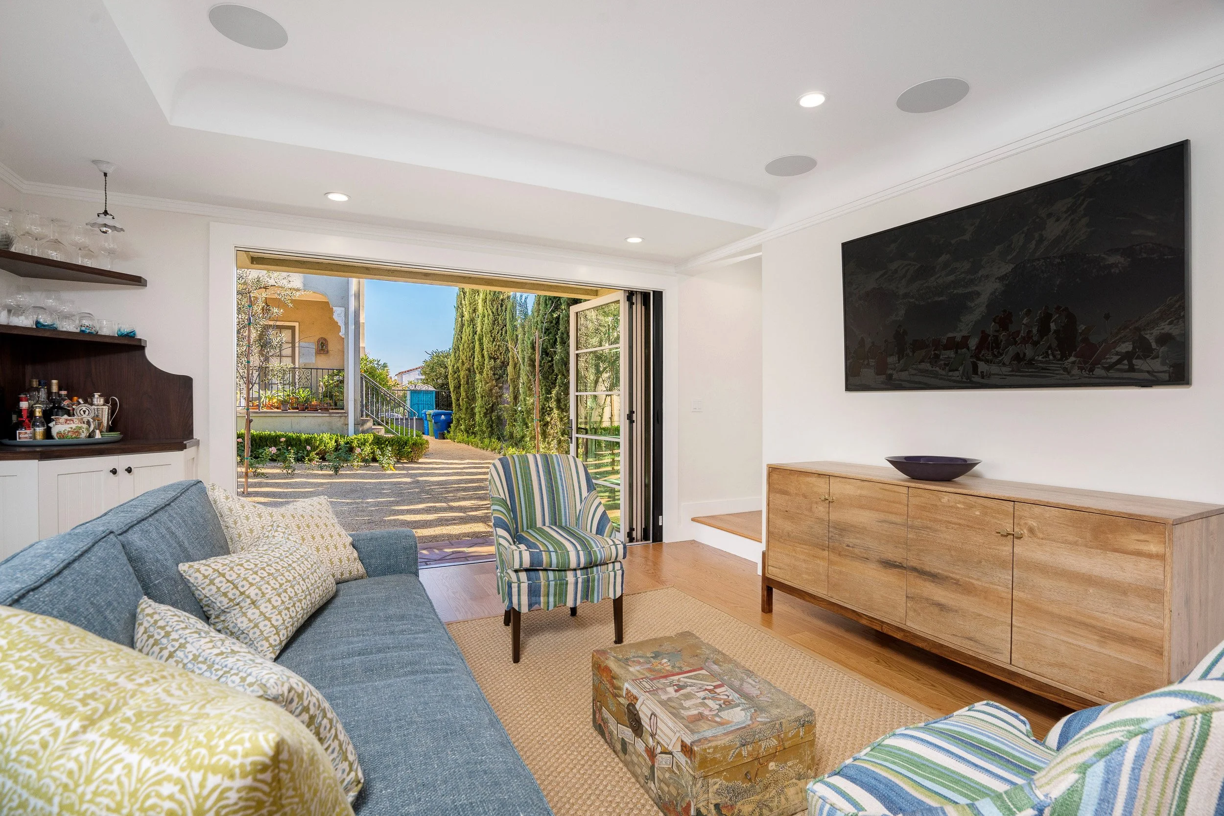 Living room with a blue sofa, striped armchair, wooden sideboard, large wall-mounted television, and open sliding door leading to a garden with trees and a blue recycling bin.