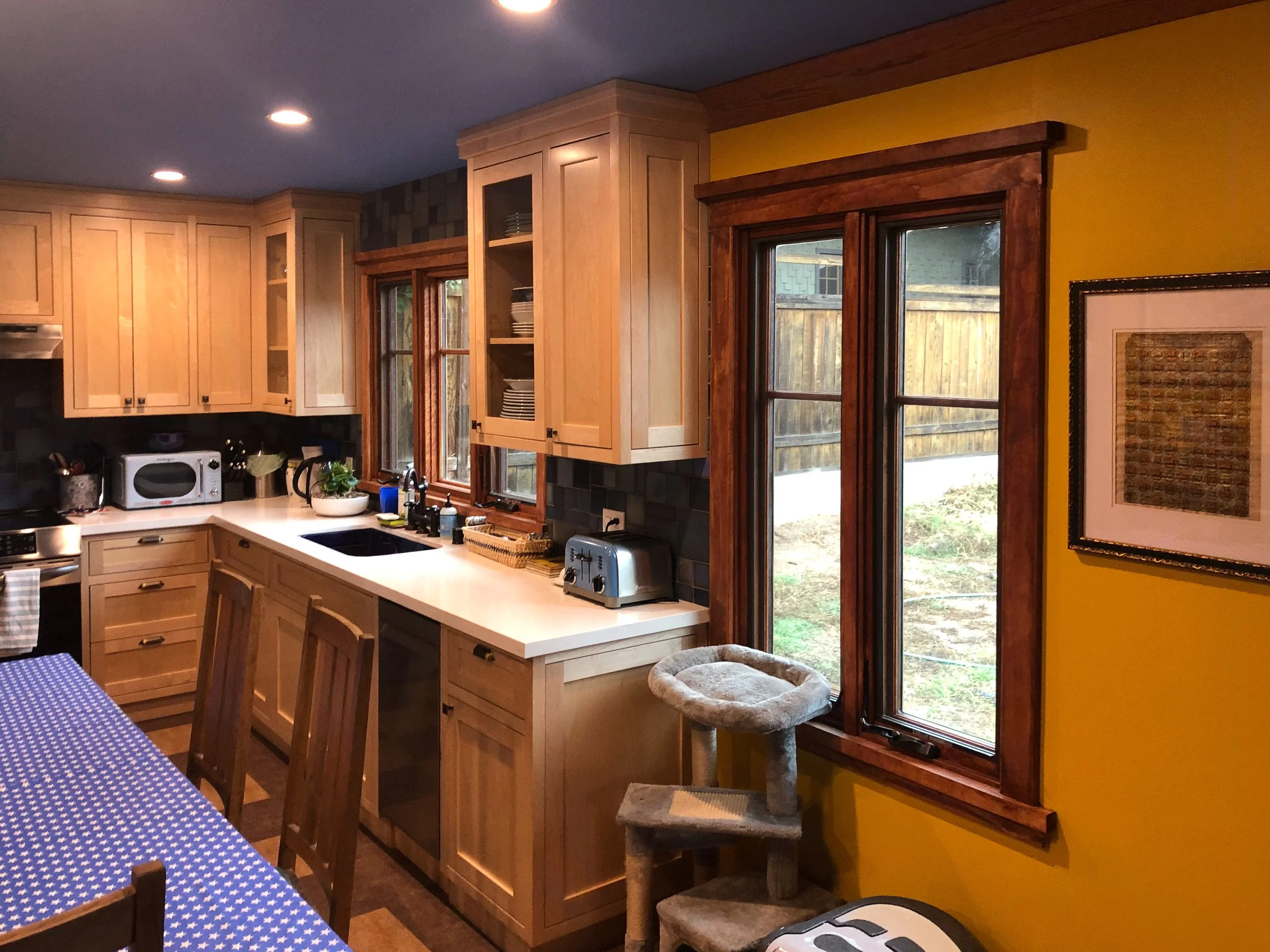 Kitchen with wooden cabinets, a yellow wall, and large windows showing a backyard.