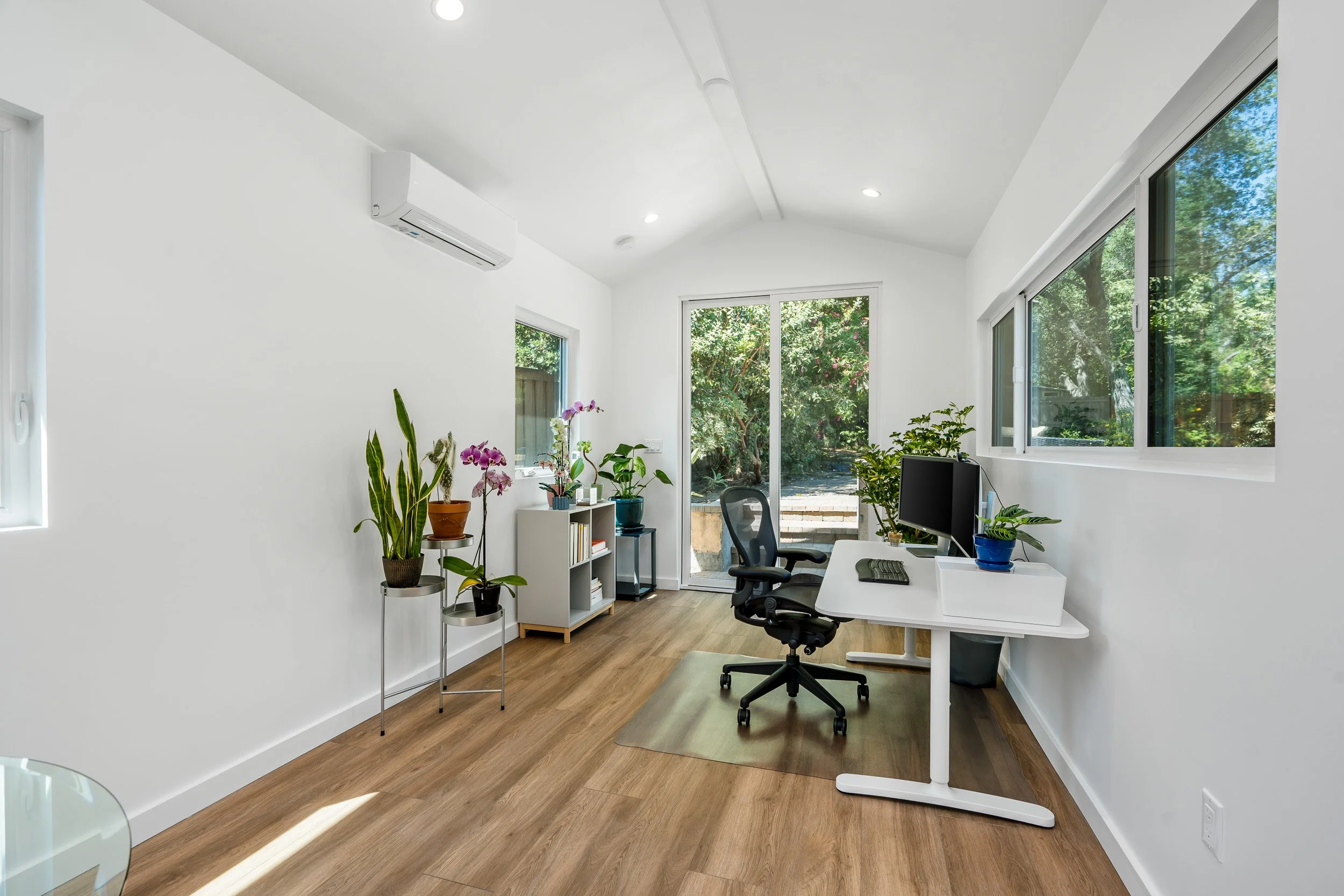 Modern home office with white walls, large windows letting in natural light, a white desk with a computer monitor, keyboard, and mouse, black ergonomic chair, and various potted plants including orchids and green leafy plants.