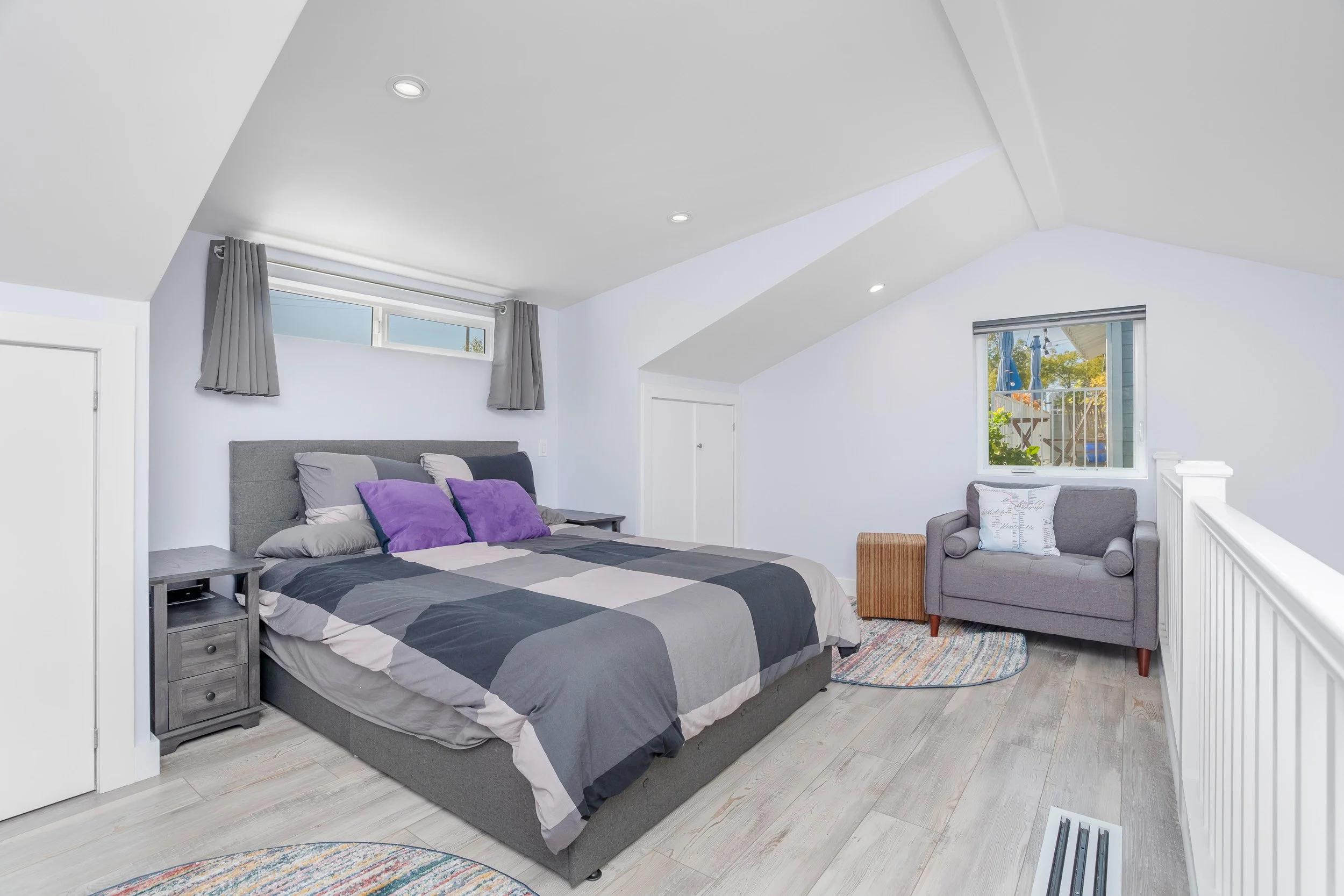 A bedroom with a gray bed frame and striped bedding, purple pillows, two gray nightstands, a gray armchair with a decorative pillow, a small wooden ottoman, and two windows with gray curtains, one of which shows a balcony with blue umbrellas outside.