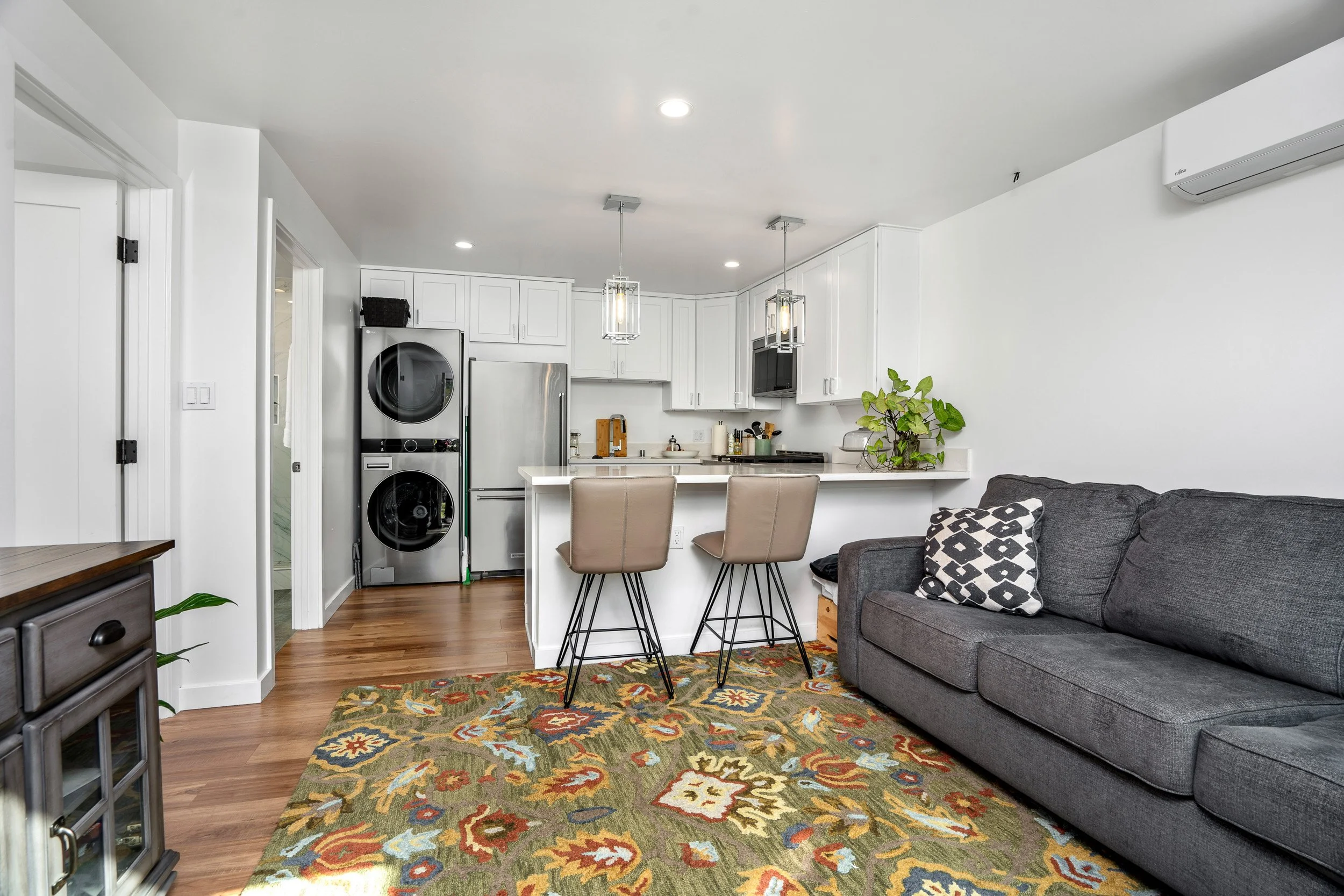 Modern living space with an open-concept kitchen and seating area. Features white cabinetry, stainless steel appliances, a gray sofa, patterned rug, and barstools at the kitchen counter.