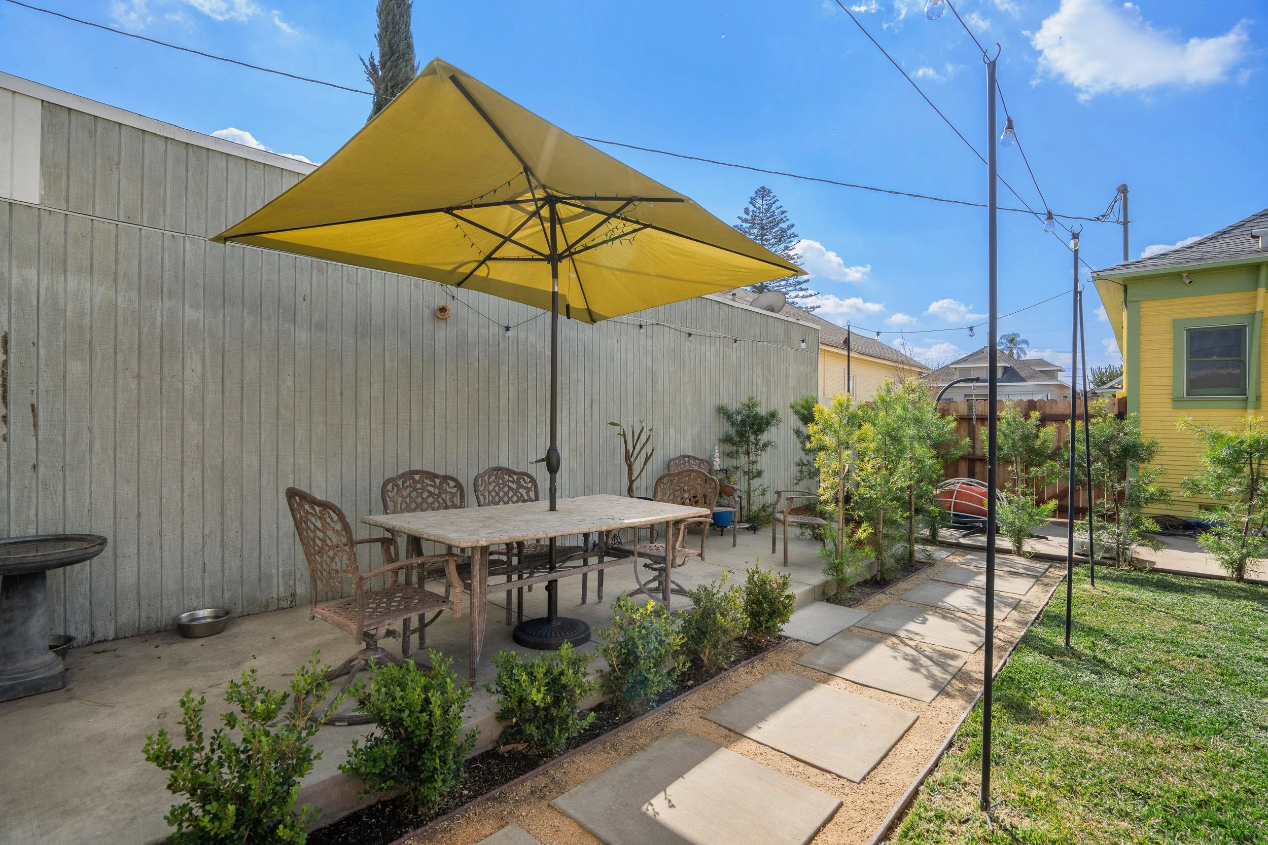 A backyard patio area featuring a yellow patio umbrella over a rectangular table with ornate chairs, surrounded by small shrubs, with a concrete path, a green house with yellow trim, and a gray wooden fence under a blue sky with clouds.