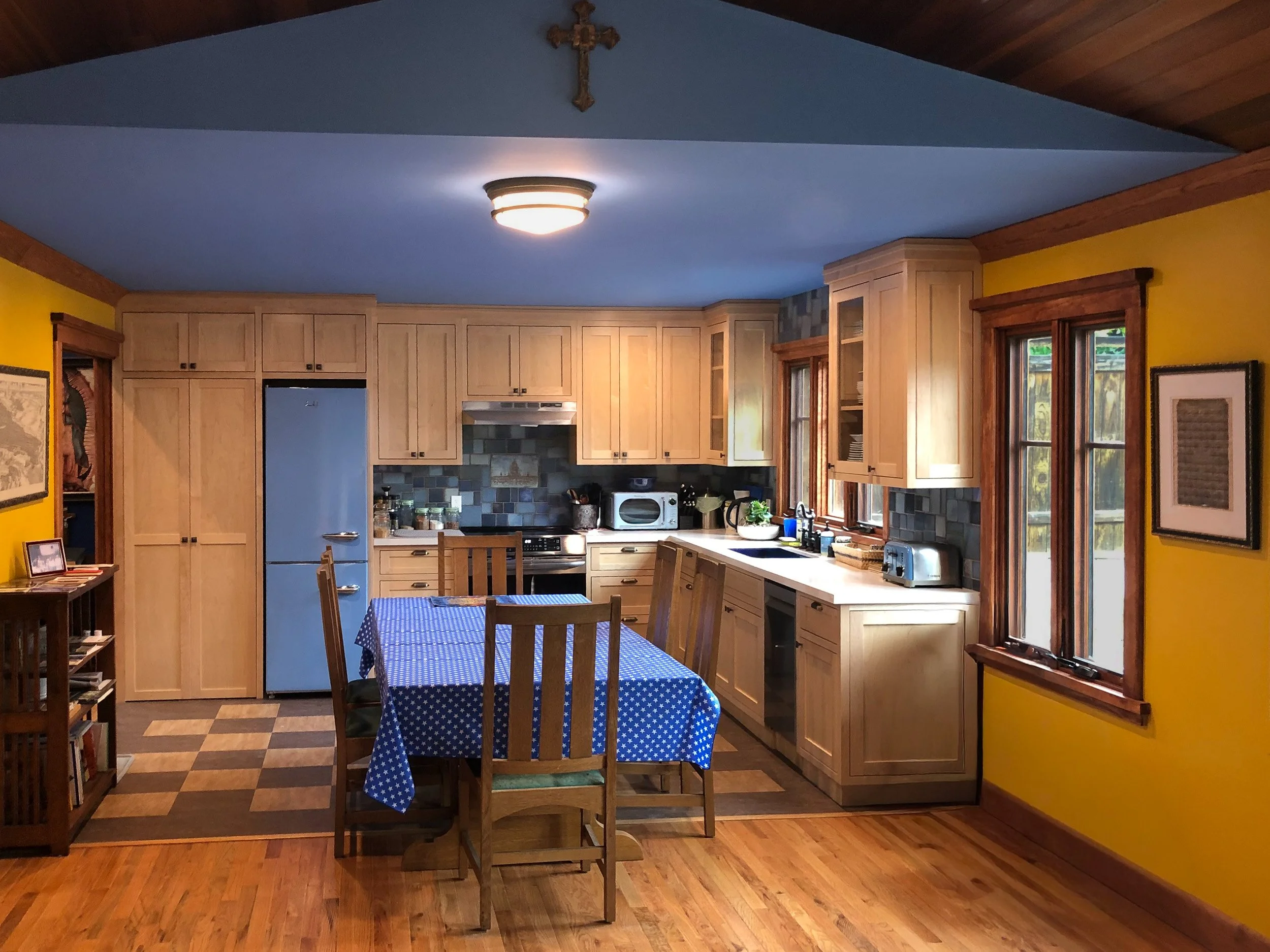 A spacious kitchen with yellow walls, wooden cabinets, a blue ceiling with a ceiling light, large windows with wooden trim, a wooden dining table covered with a blue star-patterned tablecloth, and various kitchen appliances and decor.
