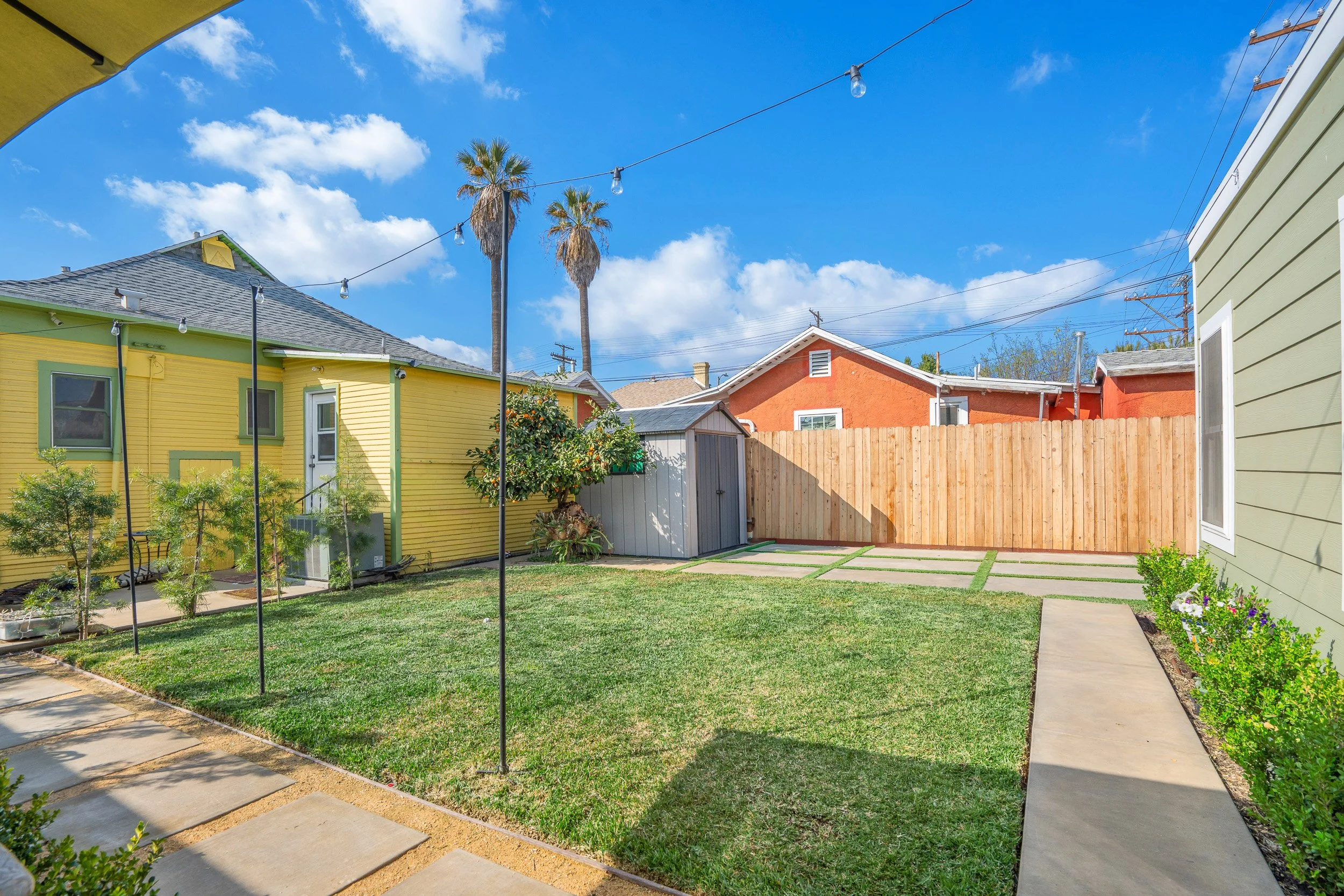 A backyard with a green lawn, concrete pavers, various plants, and wooden fences. Multiple houses with colorful exteriors, tall palm trees, and a blue sky with scattered clouds.
