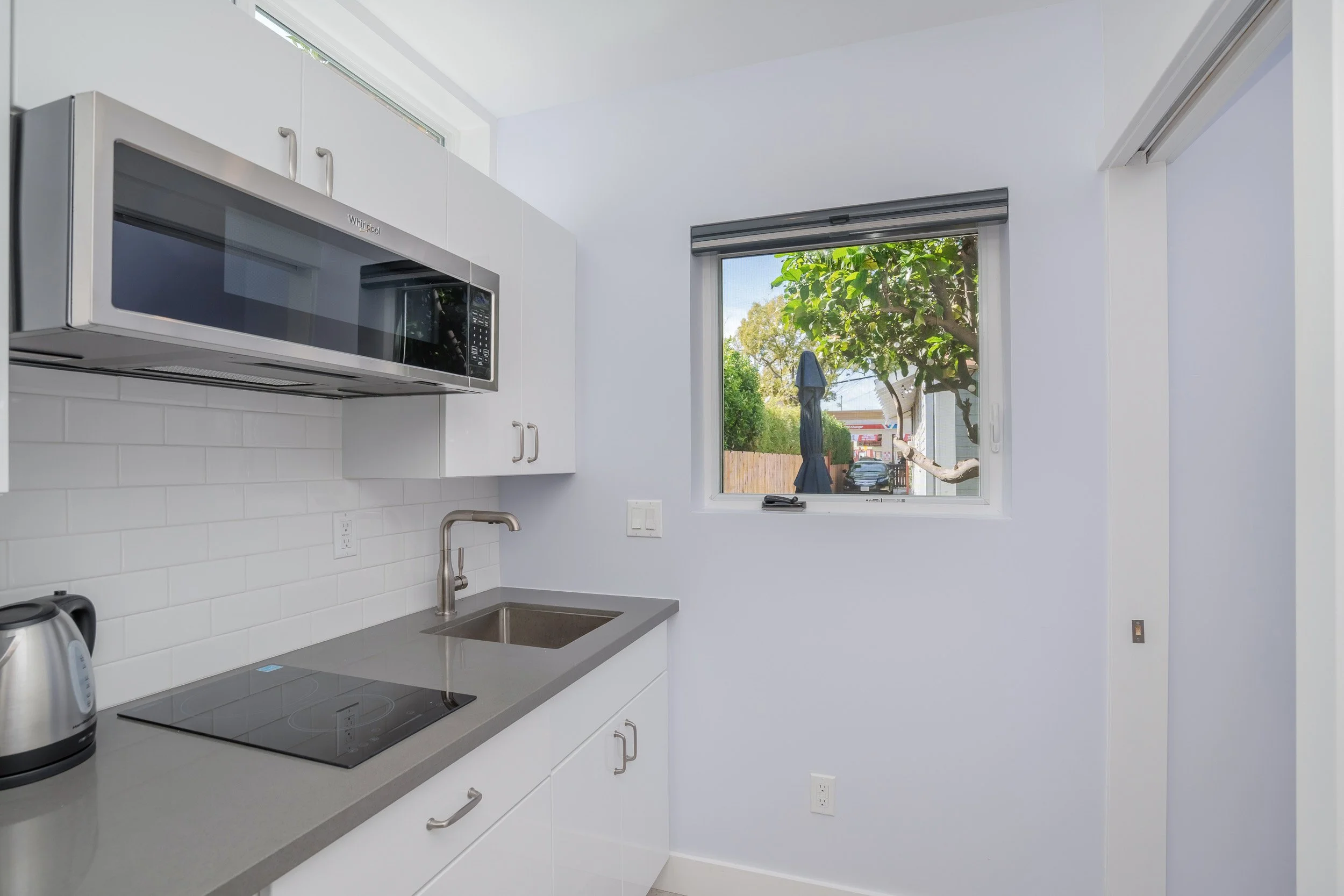 A modern kitchen with white cabinets, a gray countertop, a stainless steel microwave, a small sink, a window showing a tree outside, and a partial view of a kettle and toaster on the counter.