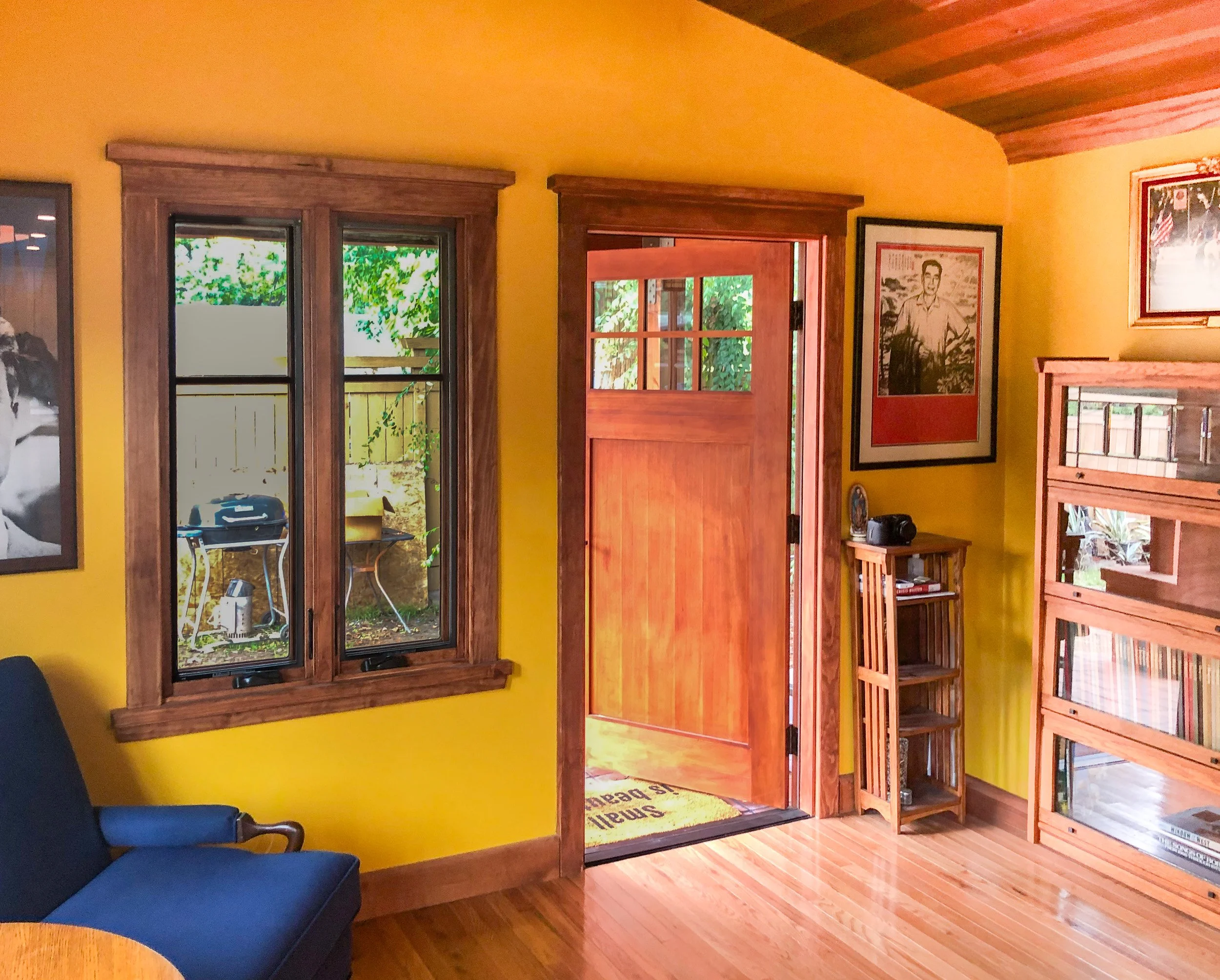 Interior of a house with bright yellow walls, wooden trim, and hardwood floors. There is a brass-framed window and a glass door open to the outside, revealing greenery. A blue cushion is on a wooden chair in the corner, and a framed black-and-white p