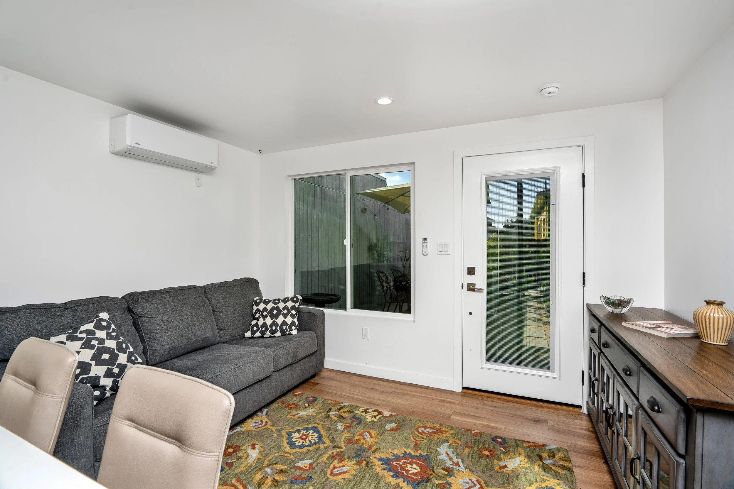 Living room with a gray sofa, patterned black and white pillows, beige armchair, colorful patterned rug, wooden sideboard with decorative items, white walls, sliding glass door with blinds, and windows showing outdoor patio with umbrella.