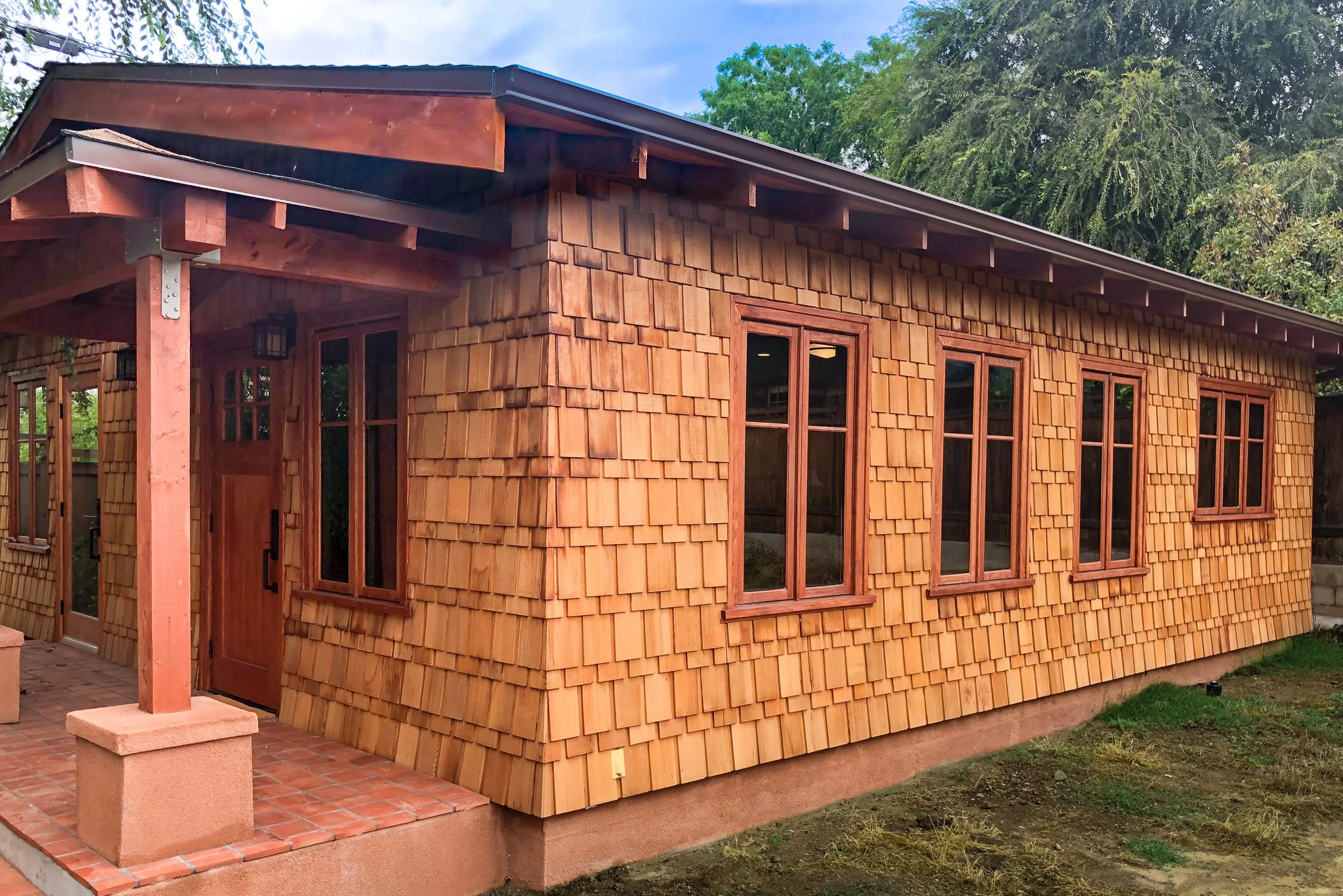 A wooden house with a shingle exterior, multiple windows, a door, and a small porch with brick flooring, surrounded by green trees.