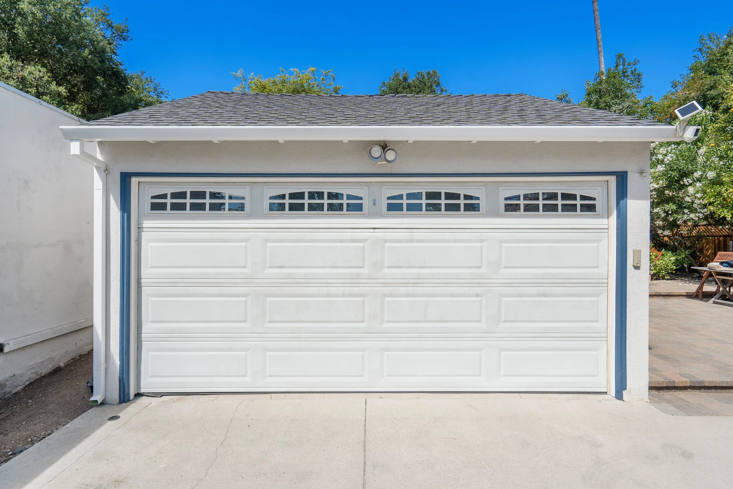 A white garage door with small rectangular windows at the top, set against the exterior of a house with a gray shingle roof. There is a circular light fixture above the garage door, a small solar panel on the right side of the roof, and green trees i
