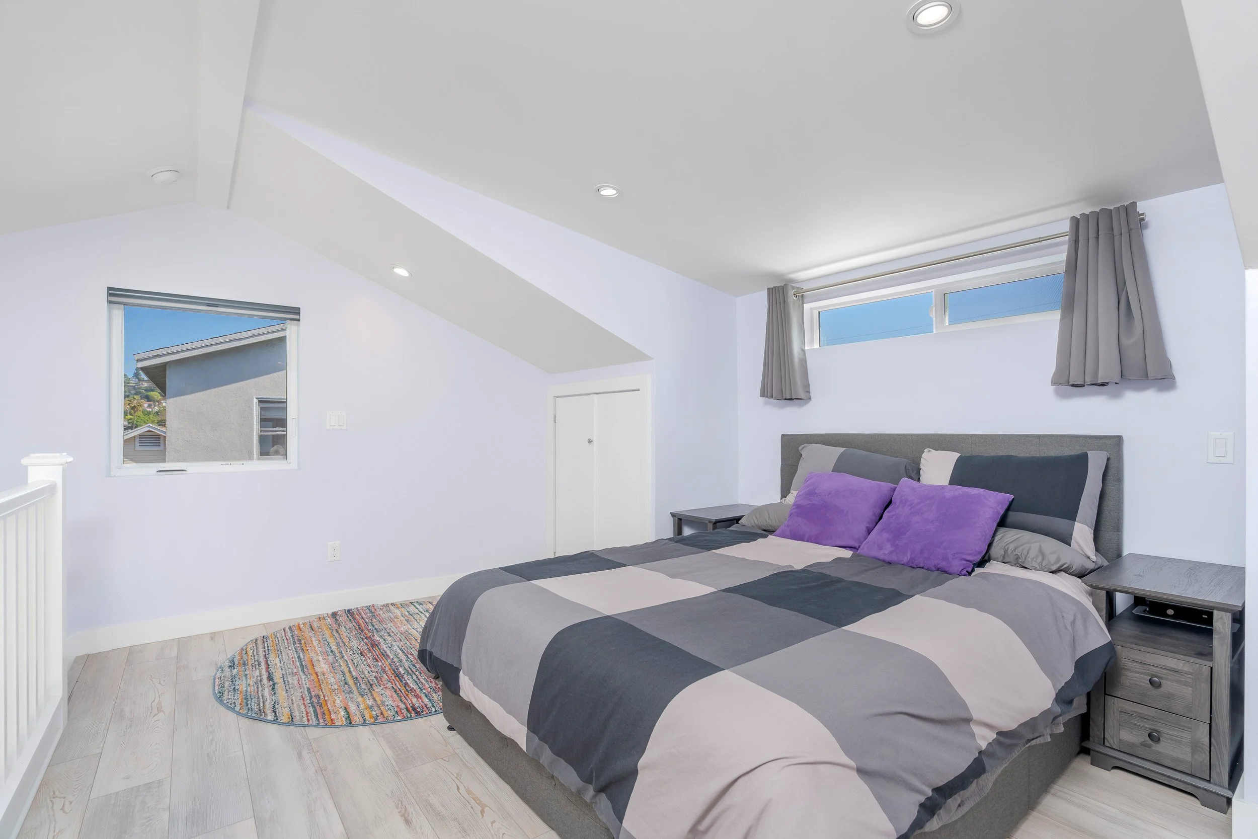 A bedroom with a gray and black striped bedspread and purple pillows. There are two gray nightstands, a window with gray curtains, and a colorful oval rug on a light wood floor.