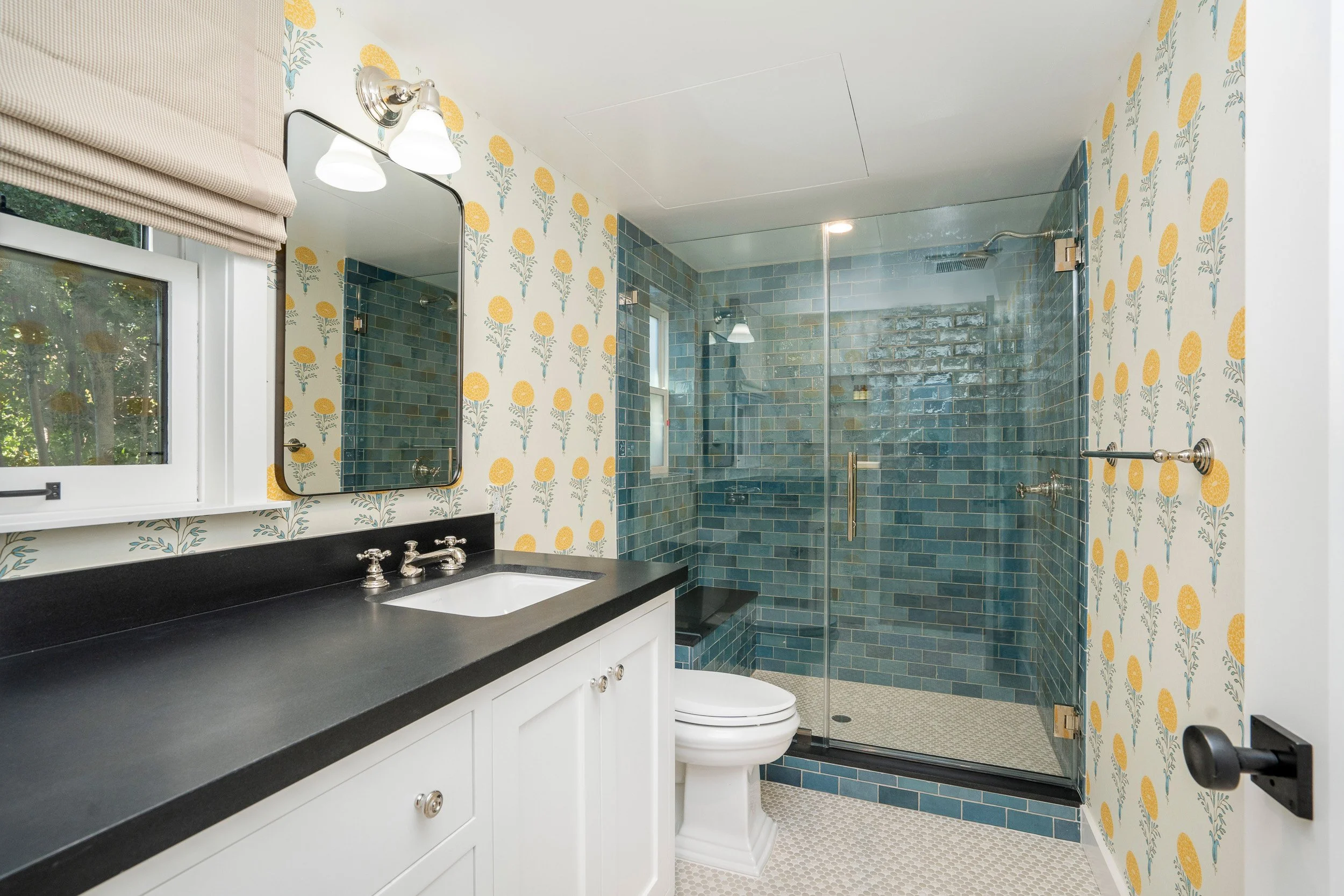 A bathroom with a vanity counter and sink, a mirror, a window with a Roman shade, floral wallpaper, and a shower with blue tile and a glass door.
