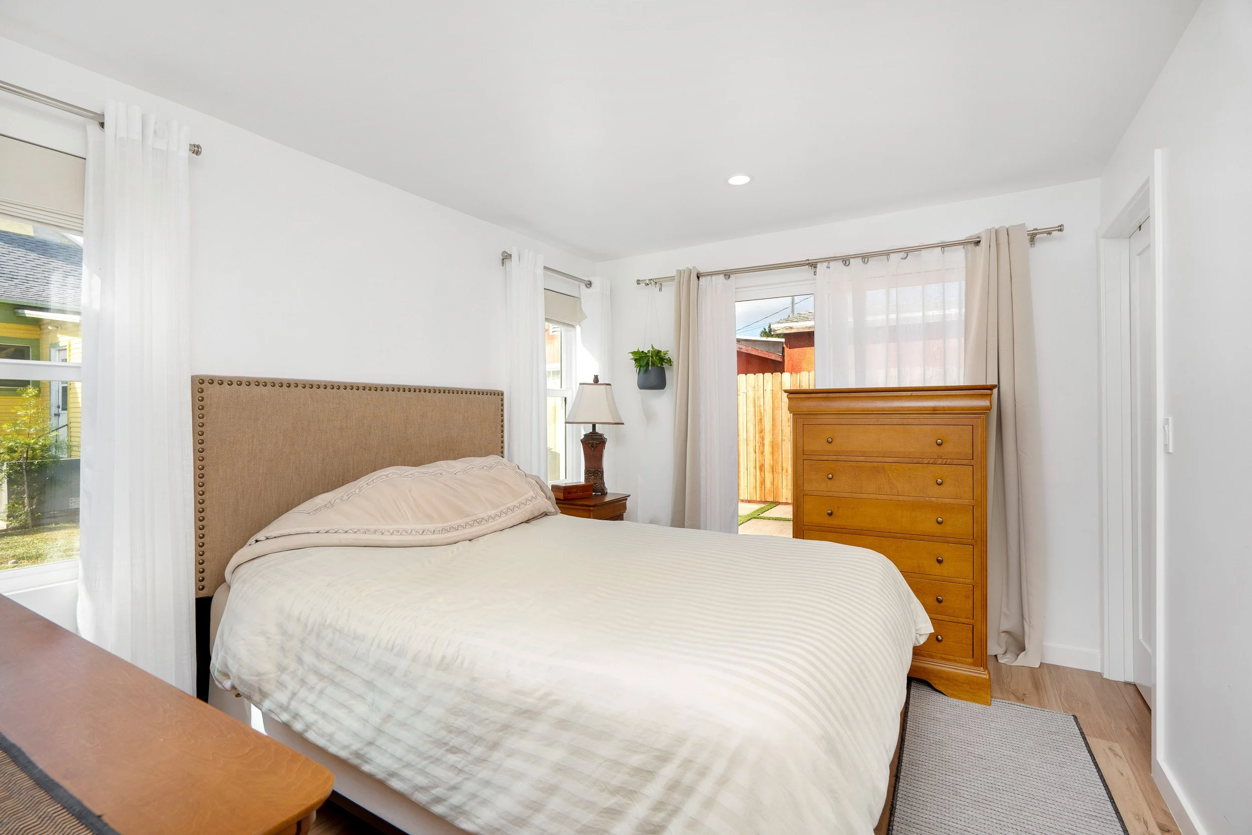 A cozy bedroom with a beige upholstered bed, white bedding, a wooden nightstand with a lamp, and a tall wooden dresser. The room has large windows with white curtains, allowing natural light to fill the space.