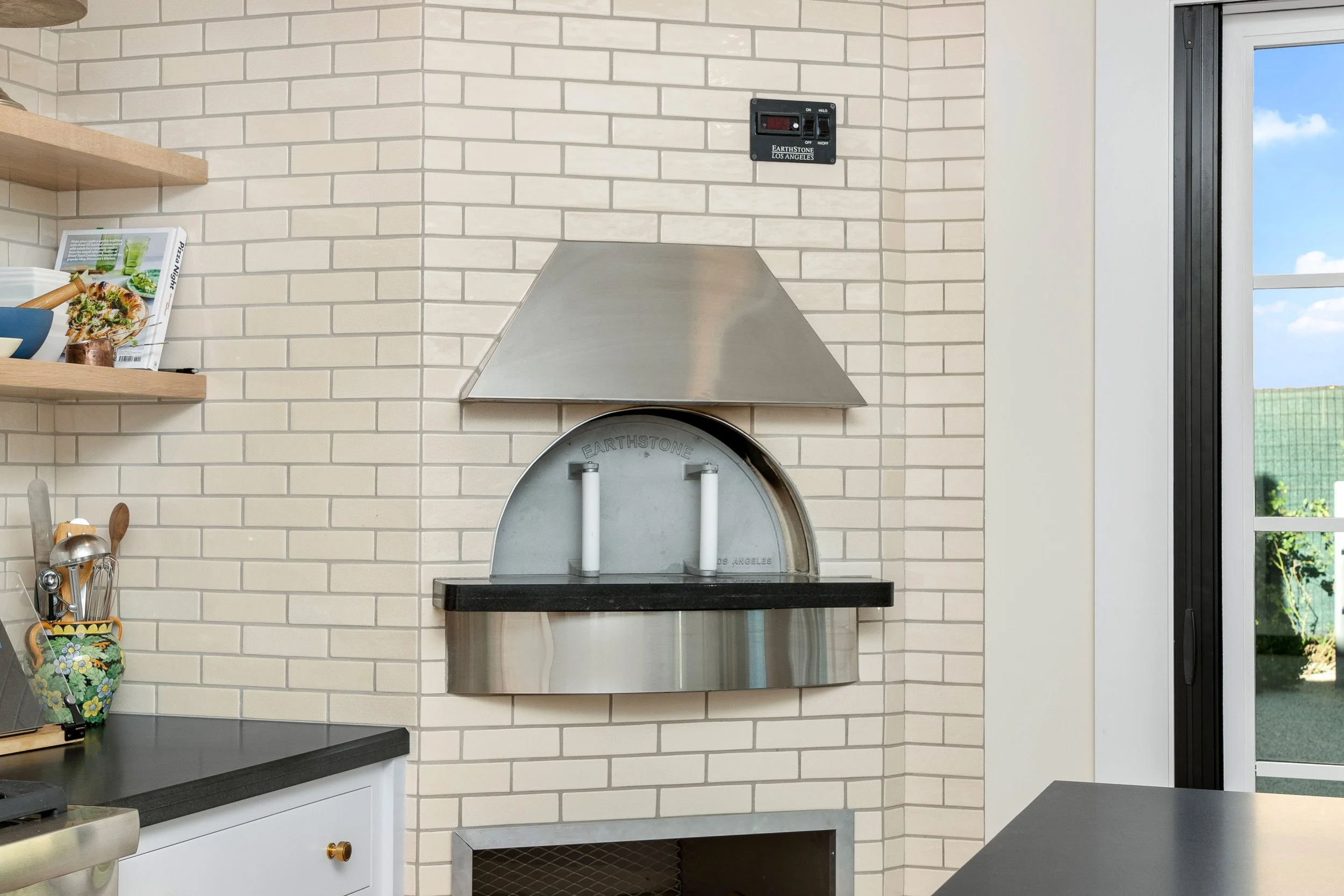 Kitchen with cream brick wall, a stainless steel outdoor pizza oven, a black countertop, white cabinetry, and a window showing a clear sky and greenery outside.