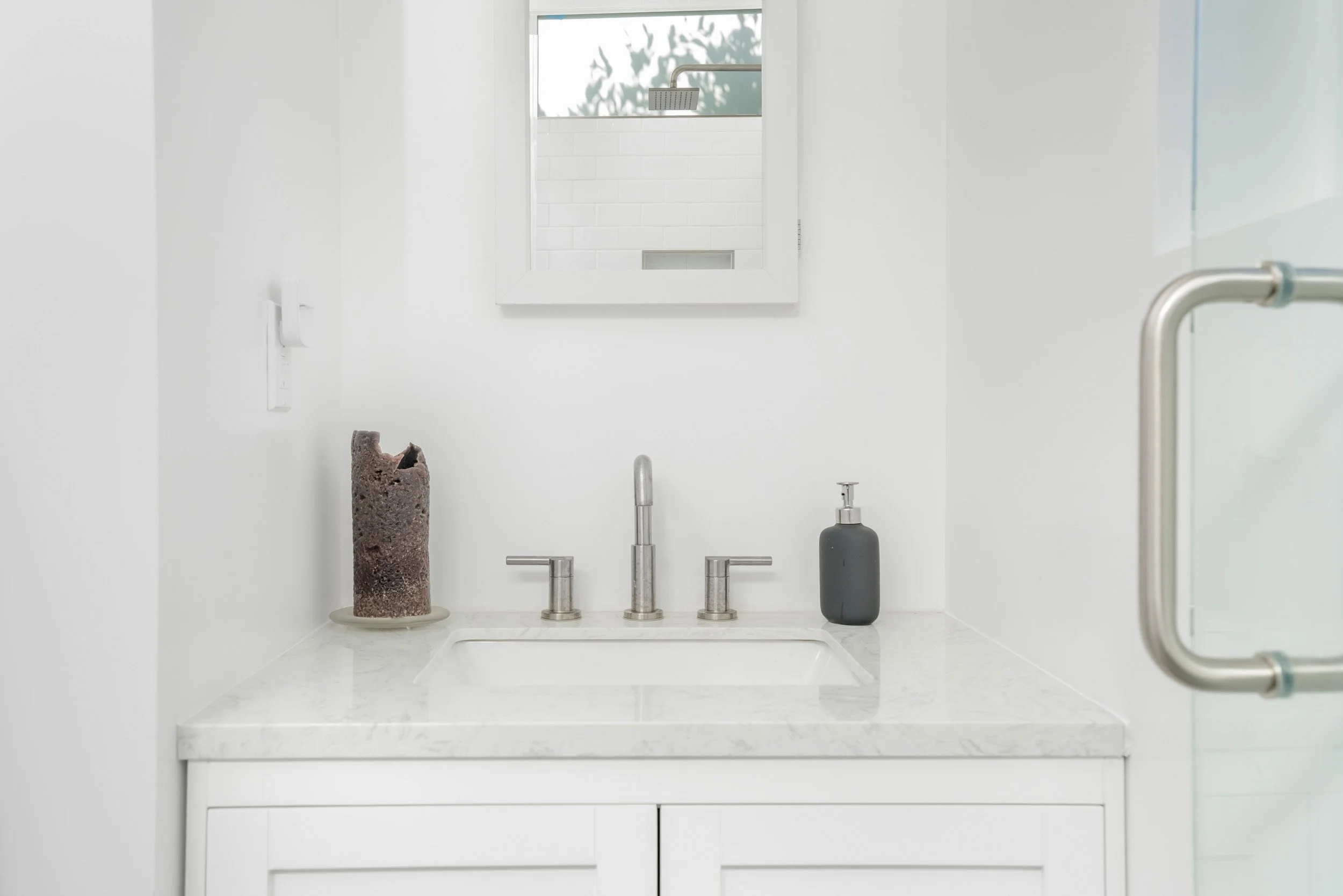 Minimalist bathroom with white walls and countertop, sink with silver faucets, a dark soap dispenser, a tall textured brown vase, and a decorative cylindrical object, with a mirror above the sink and a window in the background.