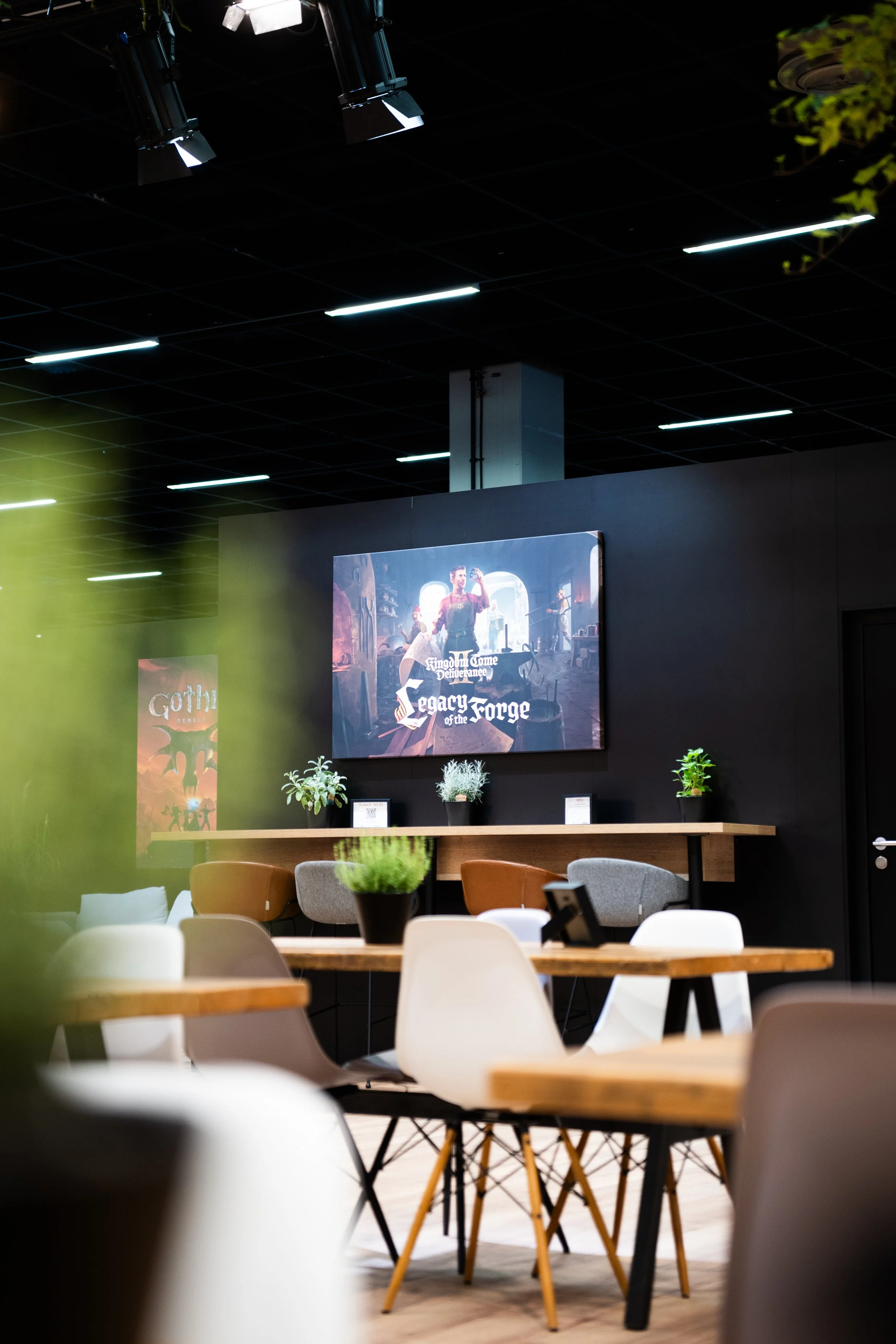 Interior of a modern communal space with wooden tables, white and gray chairs, potted plants, and framed posters on a dark wall, illuminated by ceiling lights.