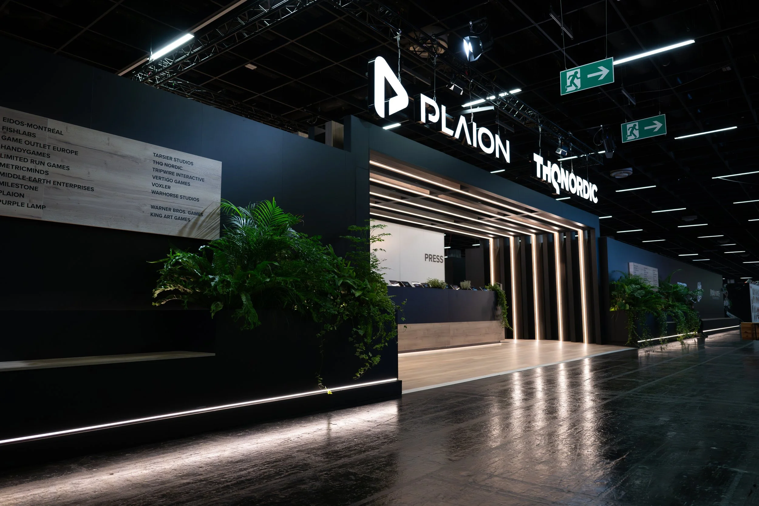 Interior of a modern exhibition hall with illuminated signs displaying 'PLAYON' and 'THNORADIC'. There are green plants and a signboard listing various game studios such as 'EIDOS-MONTREAL' and 'WARHORSE STUDIOS'. Exit signs with running figure symbols are hanging from the ceiling.
