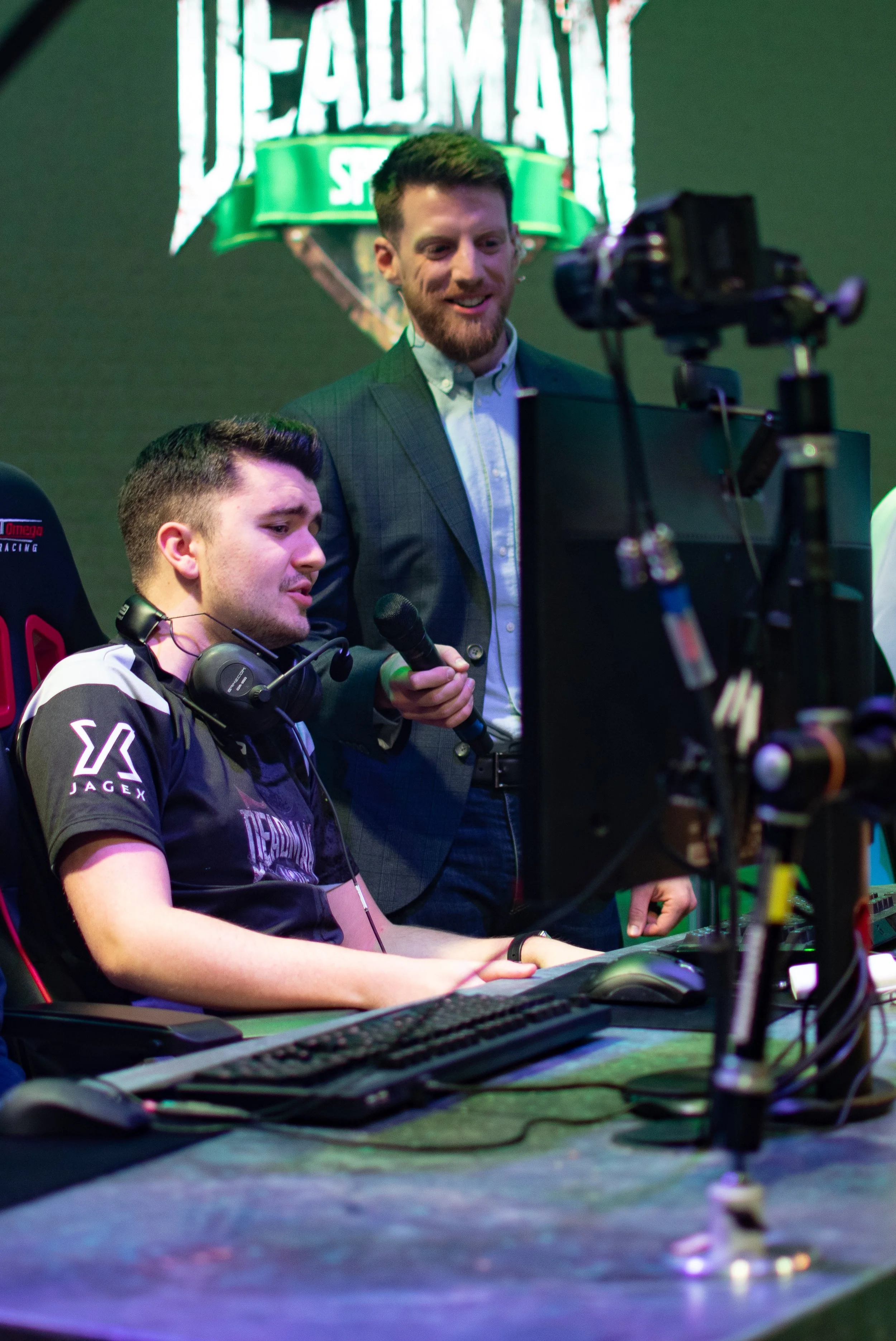 A young man sitting at a gaming station with a headset and gaming gear, and an older man standing beside him holding a microphone, both watching a computer screen. There is a blurred gaming logo in the background.