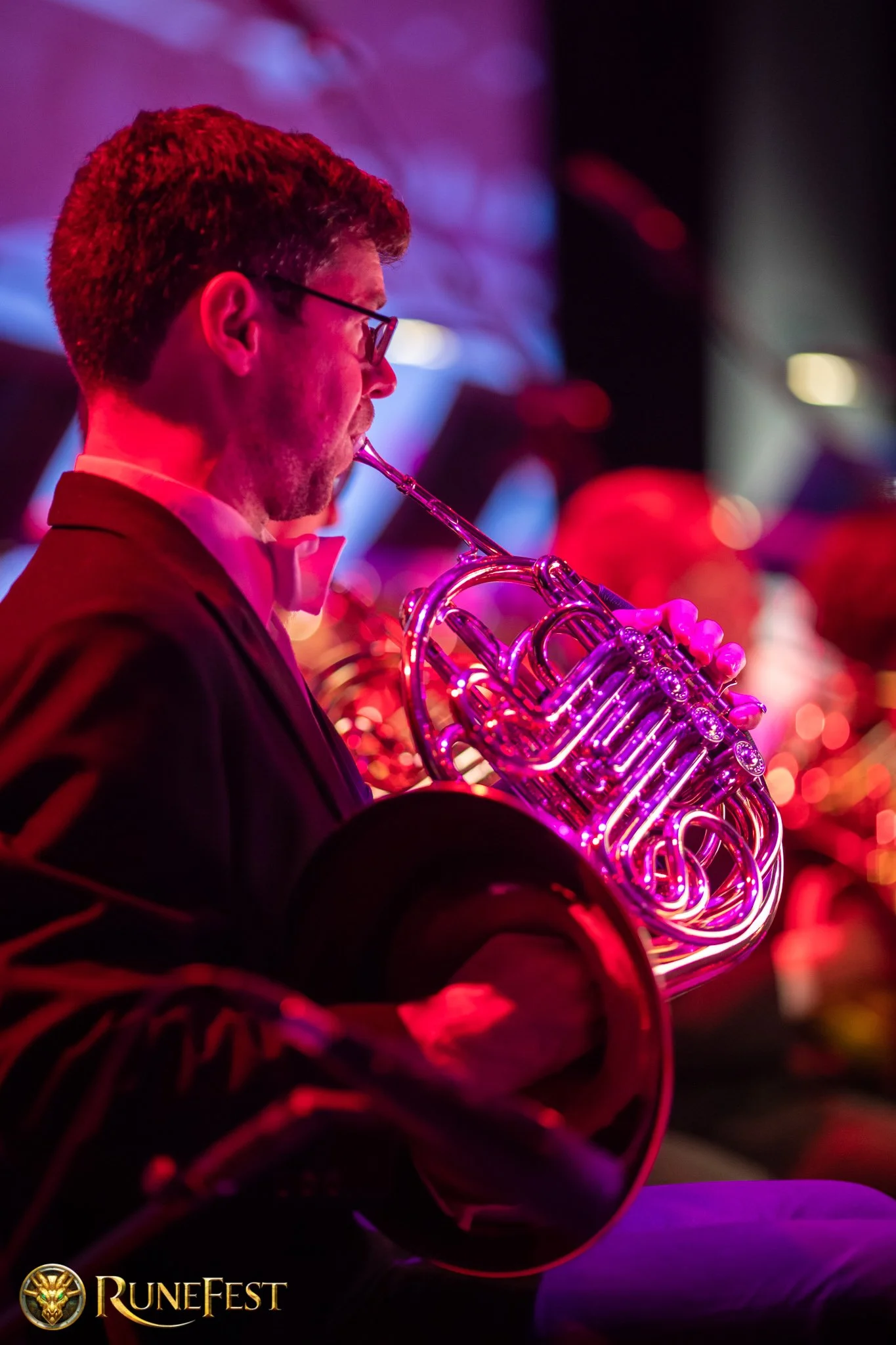 A man in formal attire, wearing glasses and a white bowtie, playing a saxophone under vibrant red and purple stage lighting at RuneFest.