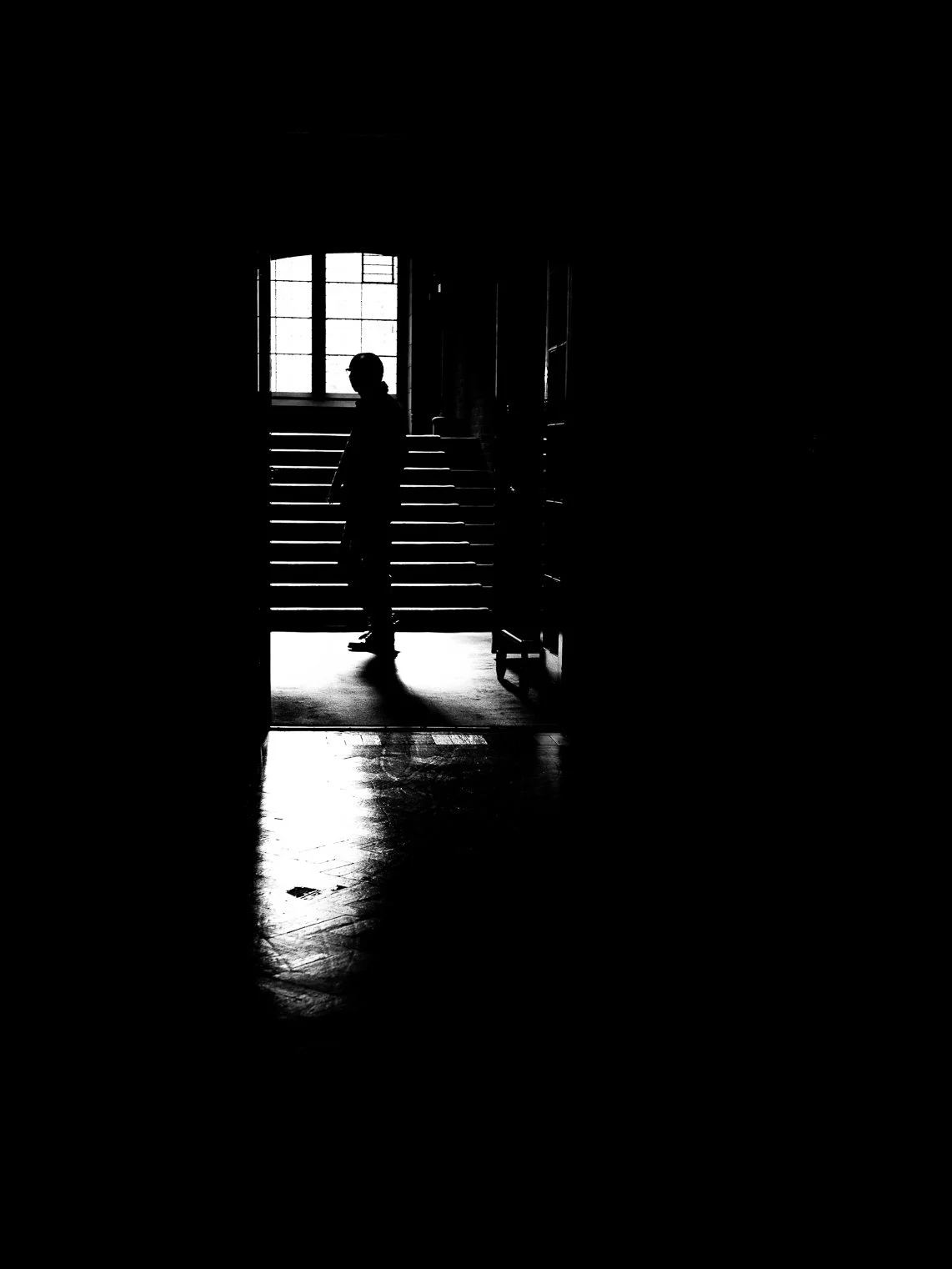 Silhouette of a person standing on stairs at the top of a doorway with a window in the background, shot in black and white.