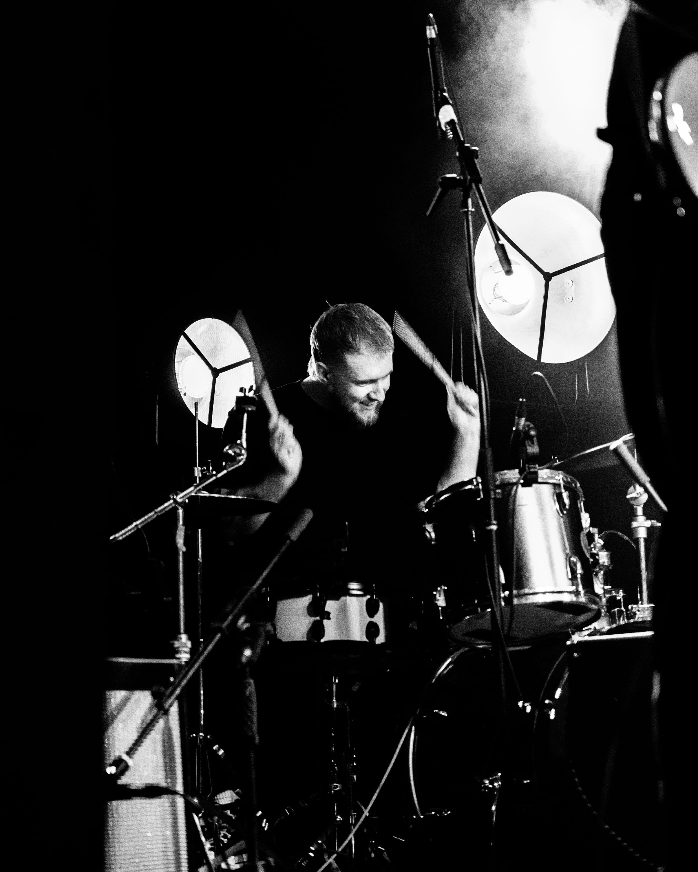 Black and white photo of a drummer smiling while playing his drum set during a performance, with stage lights and microphone stand visible.