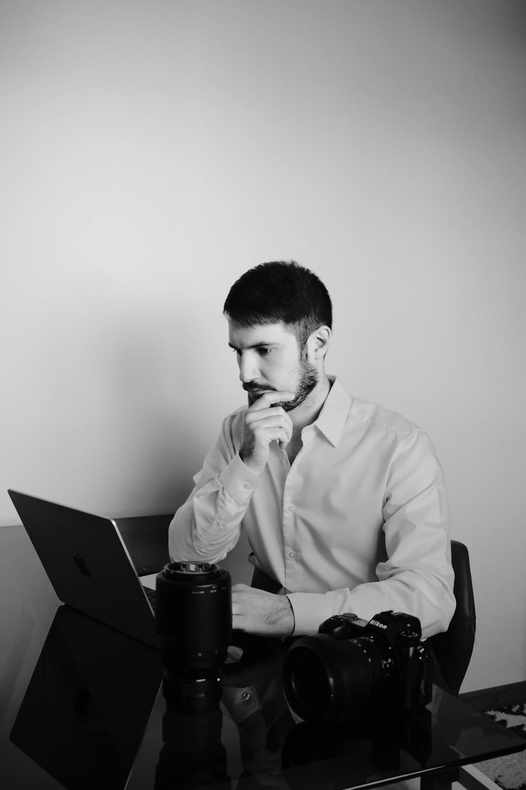 A young man with dark hair and a beard, wearing a light-colored button-up shirt, is sitting at a table with his hand on his chin, looking thoughtful at a laptop. There are professional camera equipment, including a Nikon camera and a lens, on the table.