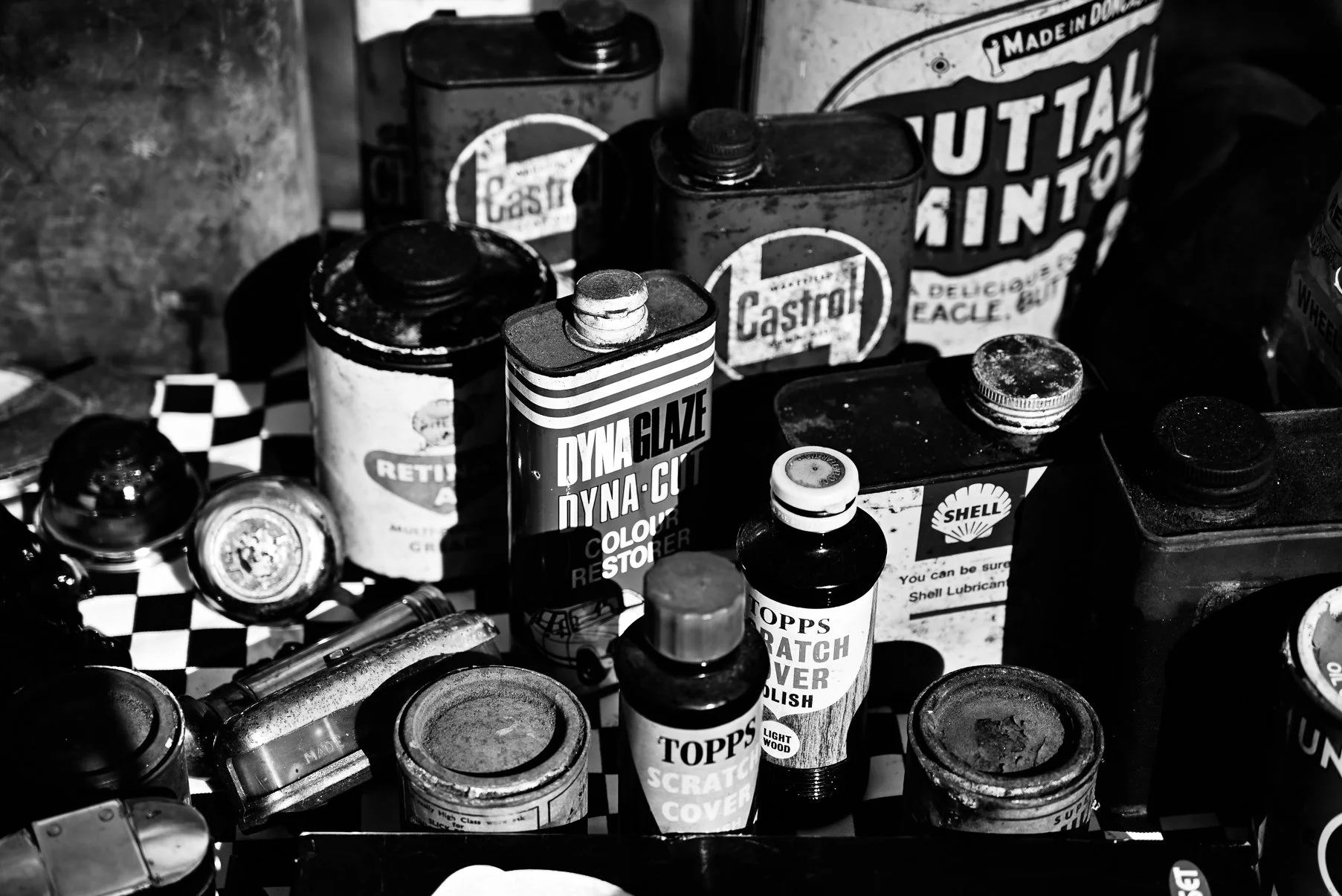 A collection of vintage oil cans, paint containers, and small bottles arranged on a checkered surface, with some labels visible including Castrol and Shell, in black and white.