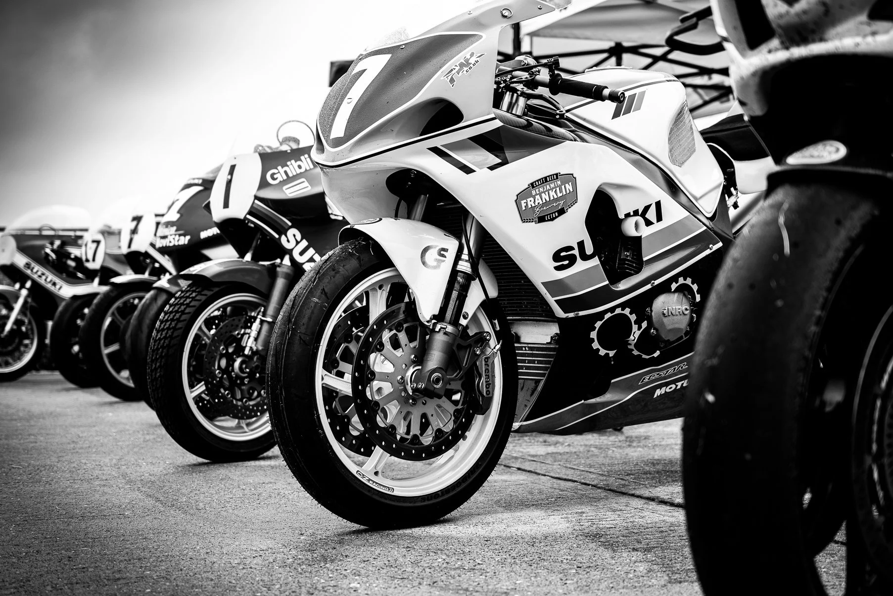 A lineup of racing motorcycles parked in a row, with the front wheels visible and motorcycles featuring various sponsor logos and number decals.