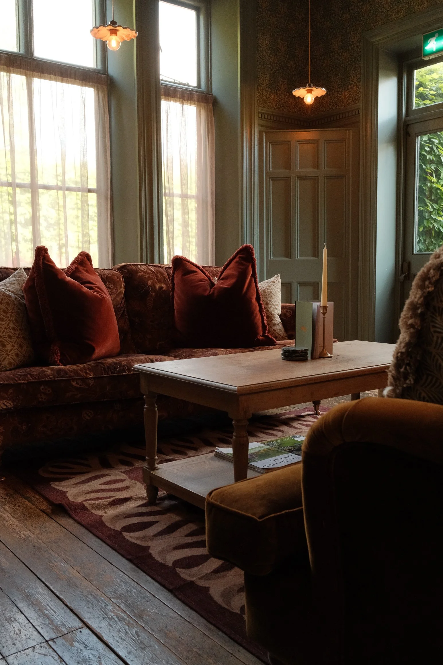 Cozy living room with large windows, red velvet cushions on a floral patterned sofa, a wooden coffee table with a candle and magazines, and warm ambient lighting from hanging lamps.