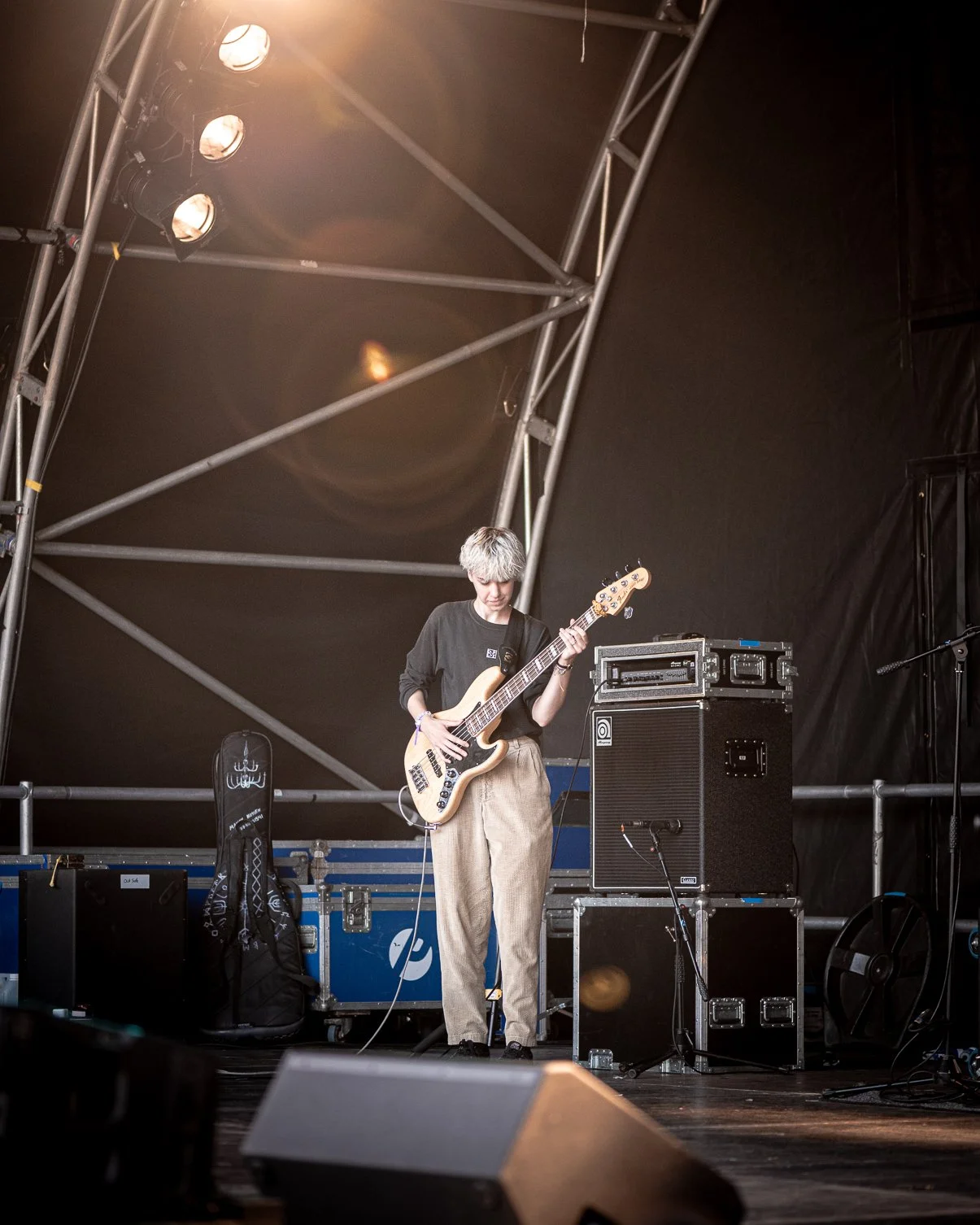 A person with short, light-colored hair playing an electric bass guitar on stage during a live performance, with stage equipment and lighting overhead.