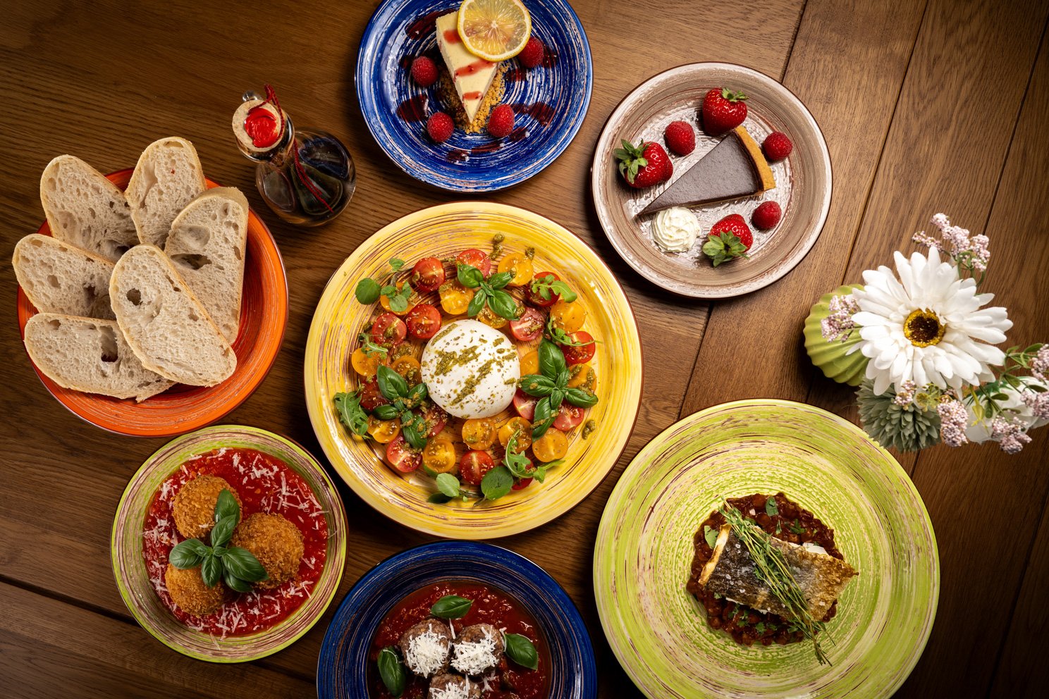 Assorted dishes including bread, salads, pizza, and desserts on a wooden table with a flower arrangement.