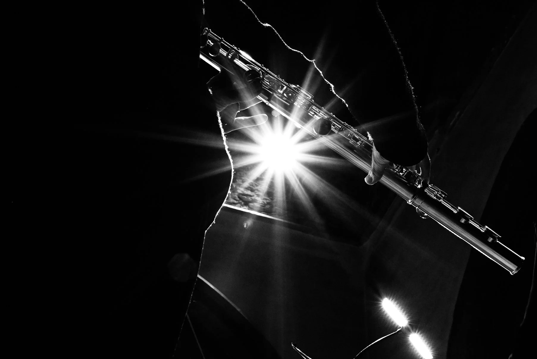 A person playing a flute or piccolo with sunlight shining behind, creating a starburst effect. The scene is in black and white with high contrast.