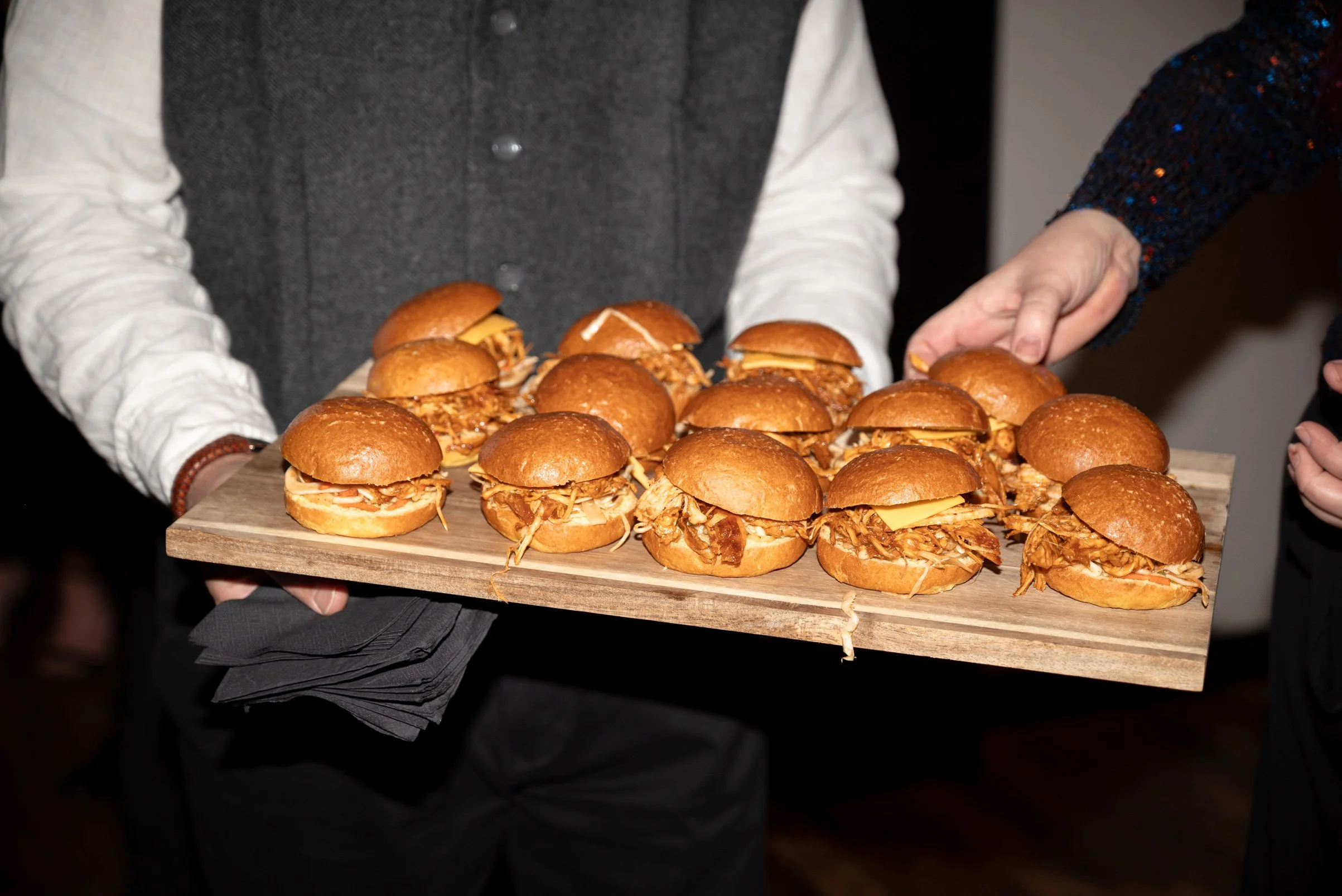 Person holding a wooden tray filled with pulled pork sandwiches with cheese on top.