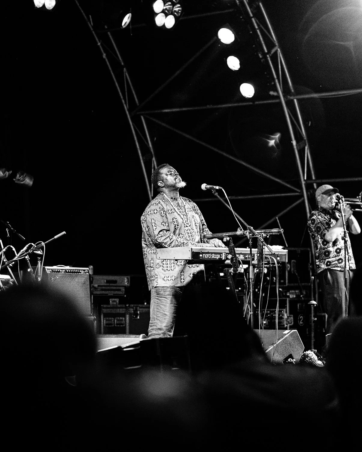 Black and white photo of a musician on stage playing a keyboard with eyes closed, and another musician singing into a microphone, with stage lights above.