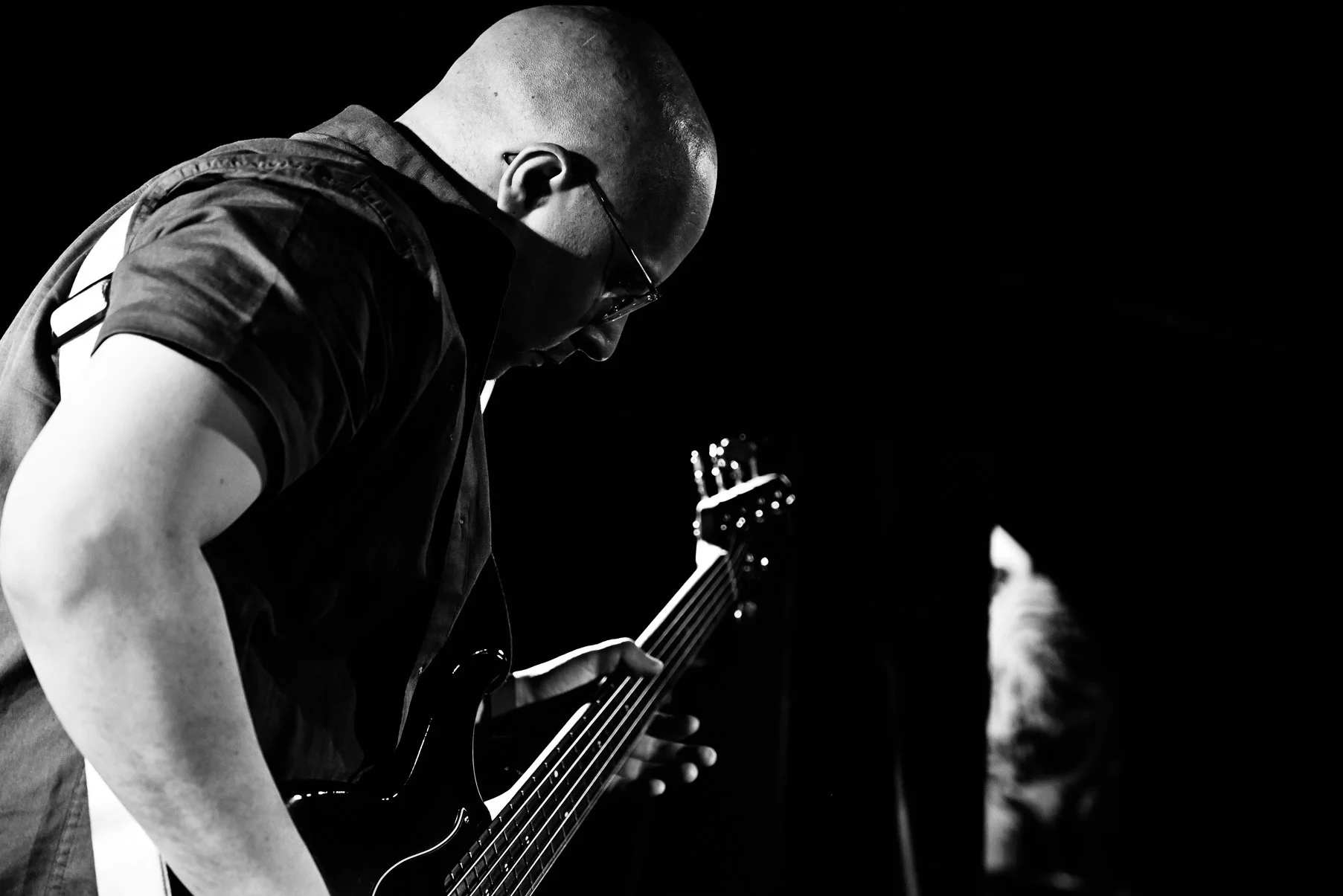 A person with a shaved head, glasses, and a tactical vest playing an electric guitar in a dark setting.