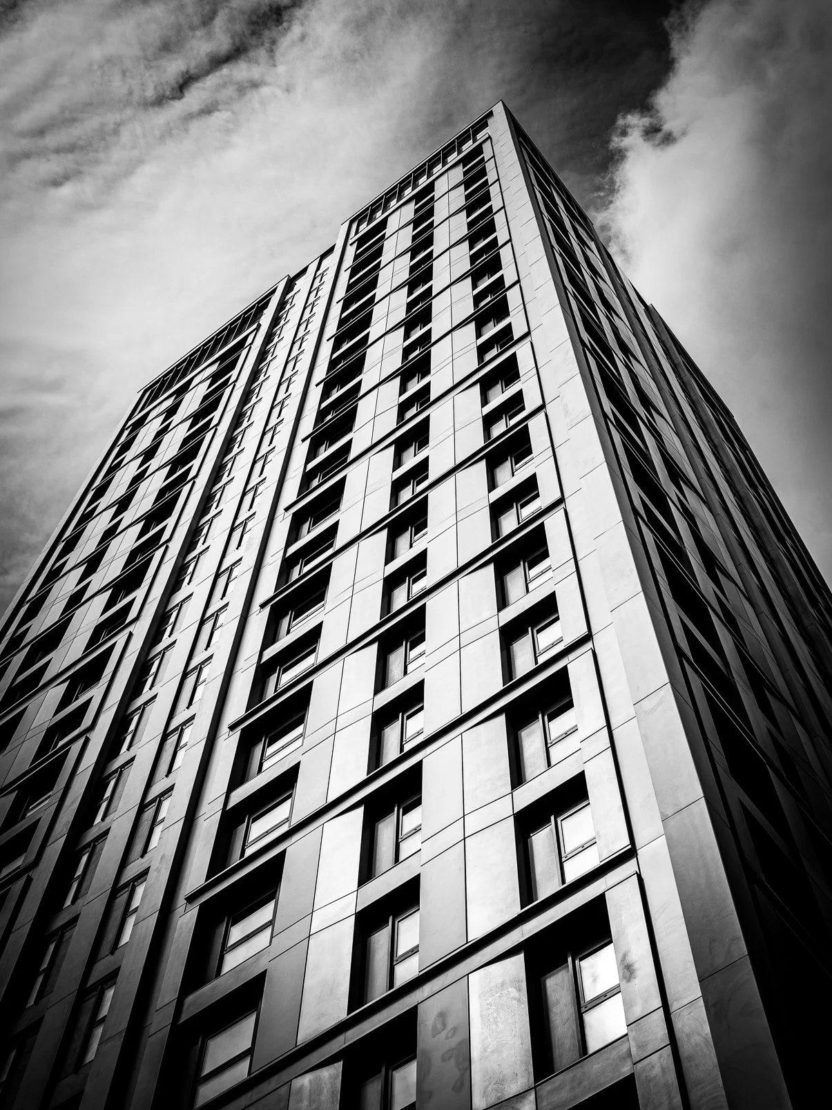 A tall modern building with a geometric design seen from below against a cloudy sky in black and white.