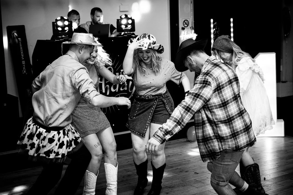 People dressed in country or Western costumes dancing at a celebration or party, with a DJ in the background.