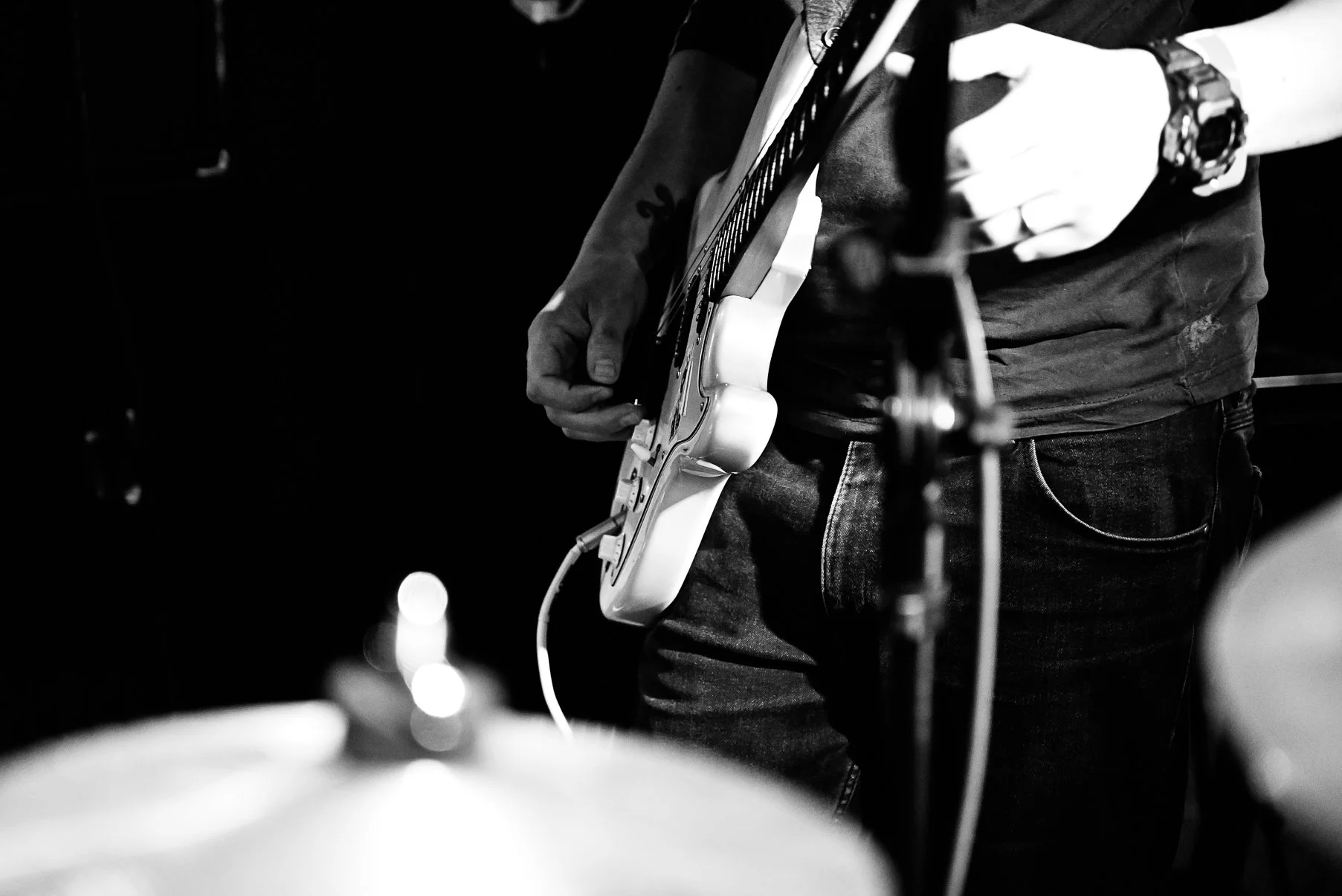 Black and white photo of a person playing an electric guitar, focusing on the torso, hands, and guitar, with only part of the face visible. The person is wearing a watch and a ring, and is standing in a dimly lit setting.