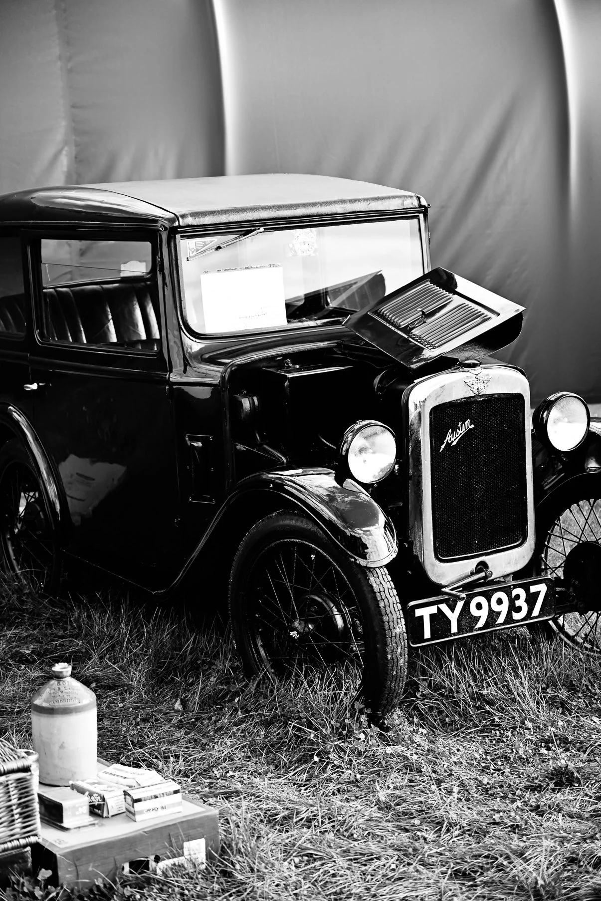 Black and white photograph of a vintage Austin car with the license plate TY 9937, parked on grass with a backdrop of a plain, fabric-covered wall. In the foreground, there are various boxes and bottles on the grass.