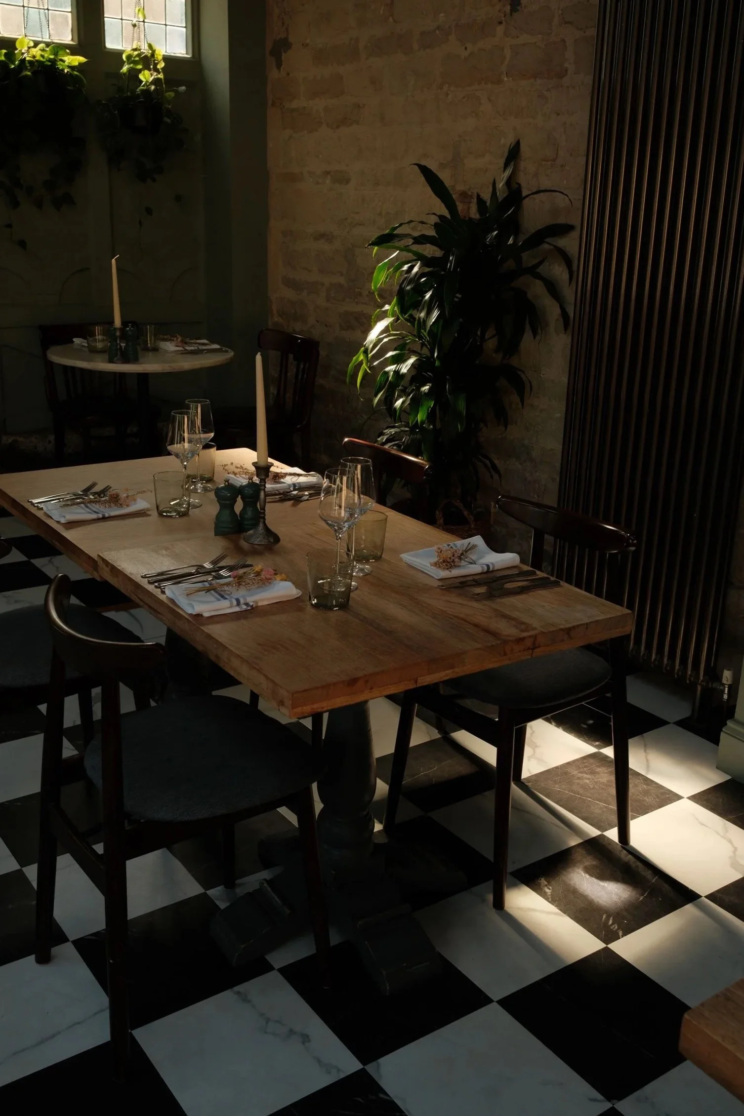 A dining room with a wooden table set with wine glasses, napkins, and candles, surrounded by chairs, and decorated with potted plants. The background features a brick wall, a window with sunlight, and a wooden partition, with a black and white checkered marble floor.