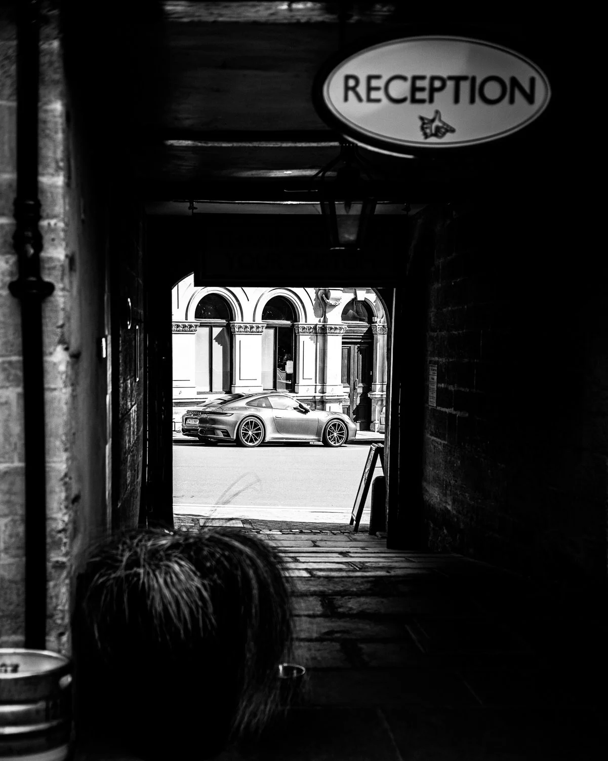 Black and white photo of a passageway with a view of a modern car parked on the street outside an ornate building with arched windows.