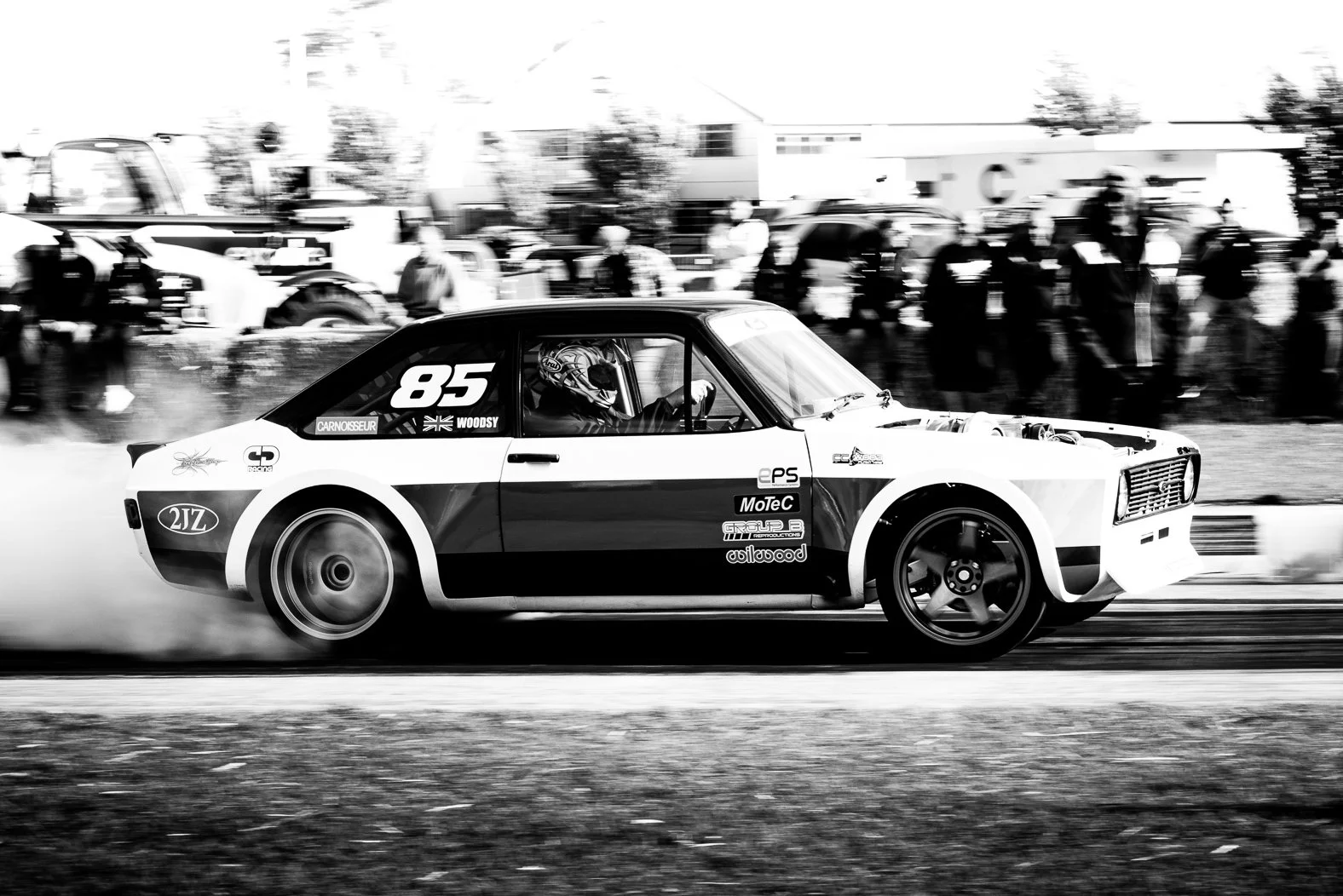 Black and white photo of a race car drifting on track with spectators and vehicles in background.