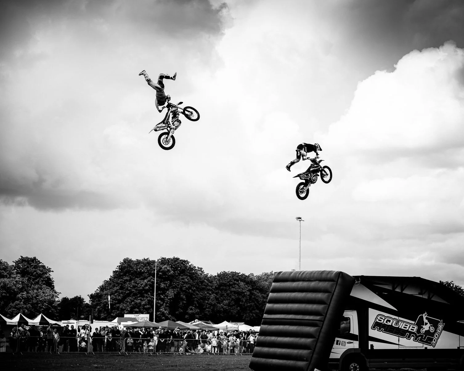 Two motocross riders performing mid-air jumps during a stunt show at an outdoor event, with spectators watching from behind a barrier.
