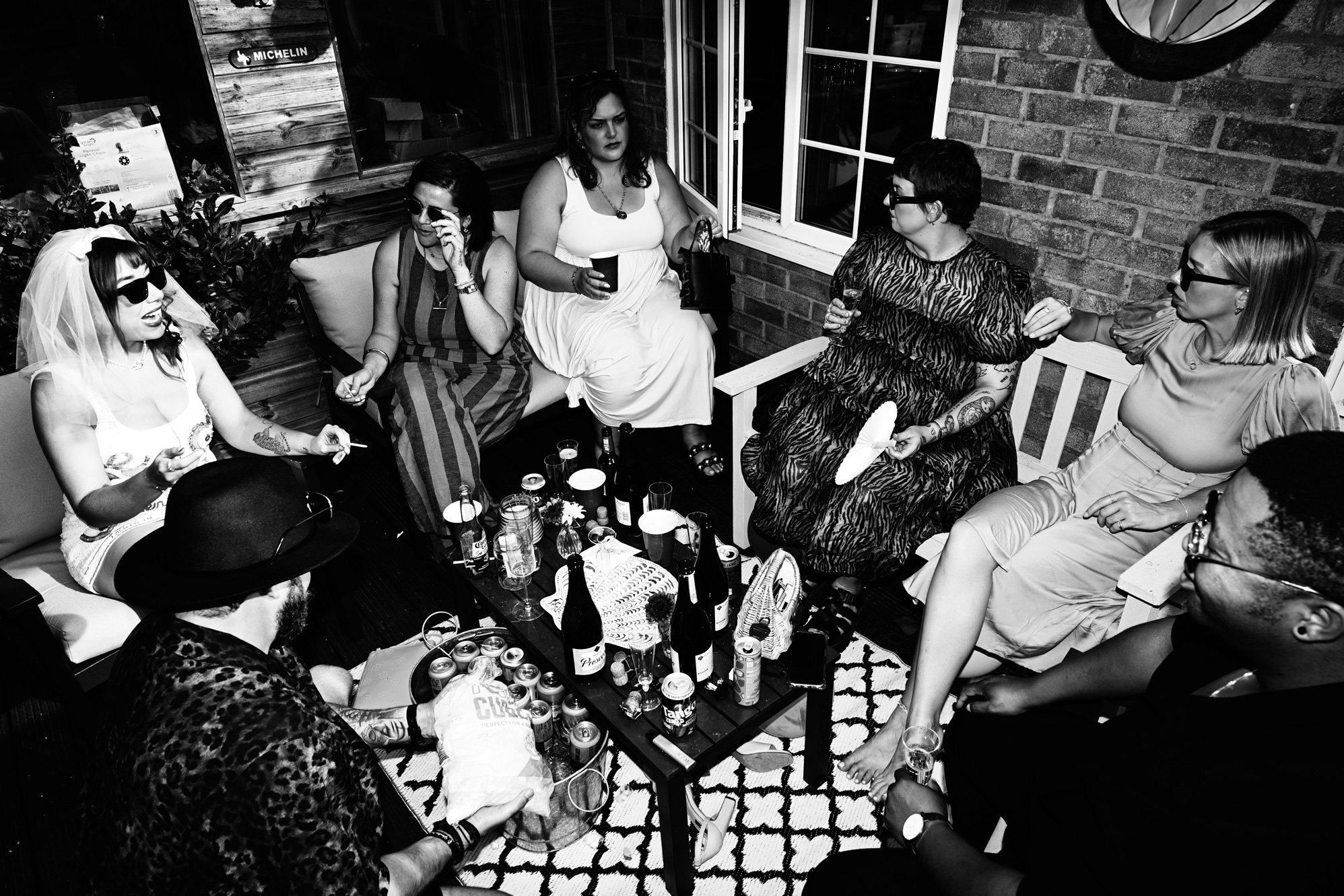A black and white photo of a group of women gathered indoors, sitting on sofas and benches, socializing with drinks and snacks on a coffee table, in a cozy setting with brick walls and windows.