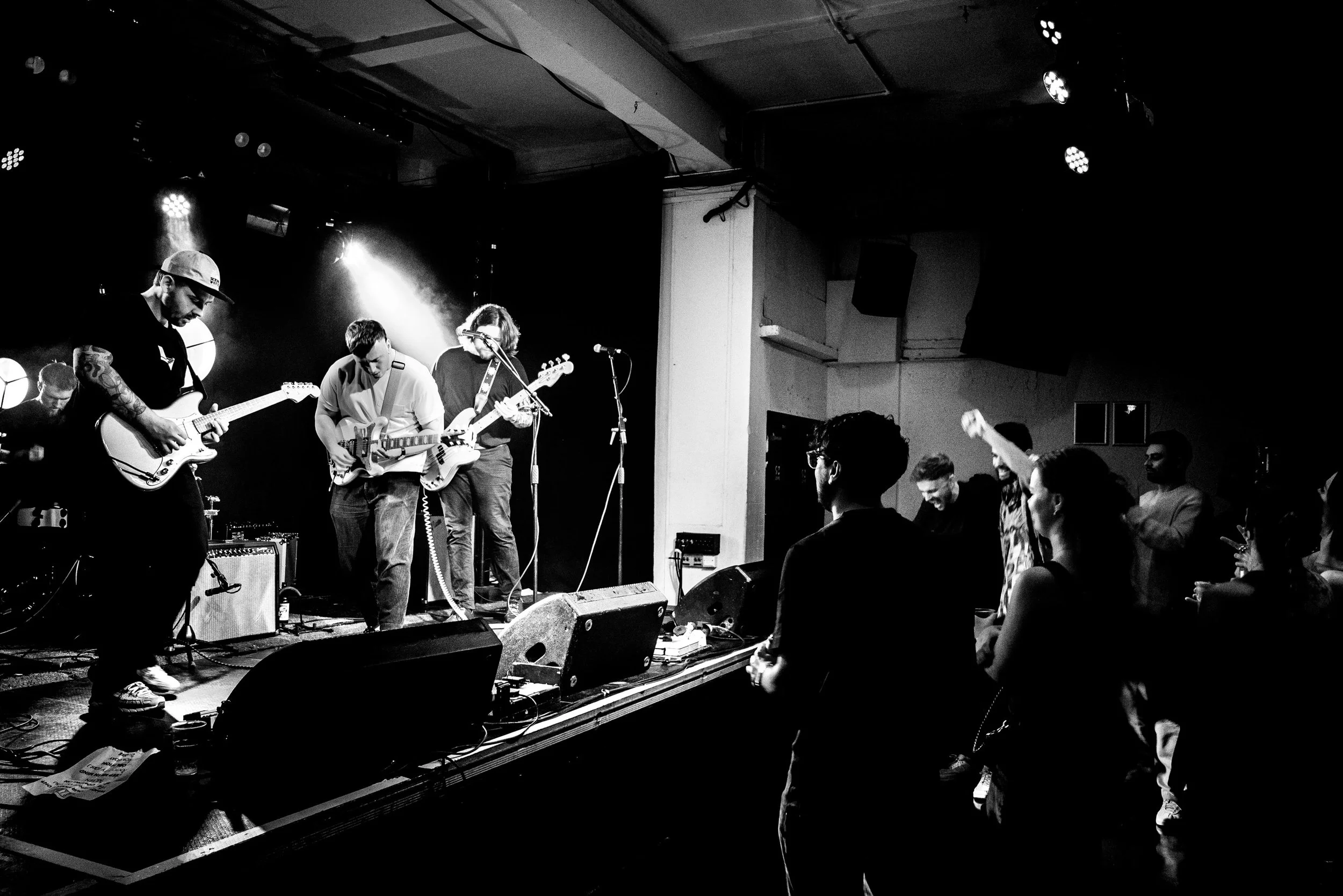 A live music performance on a stage with three guitarists playing and an audience watching, in a dimly lit venue.